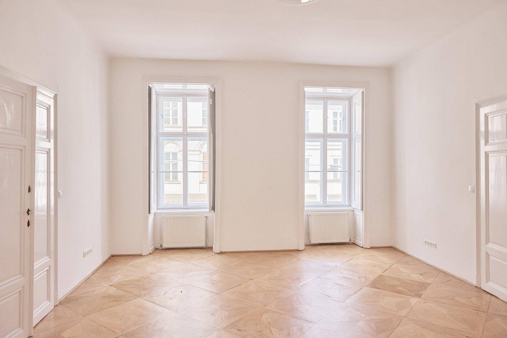 Geräumiger, heller Raum mit zwei hohen Fenstern, Parkettboden und einer Tür.