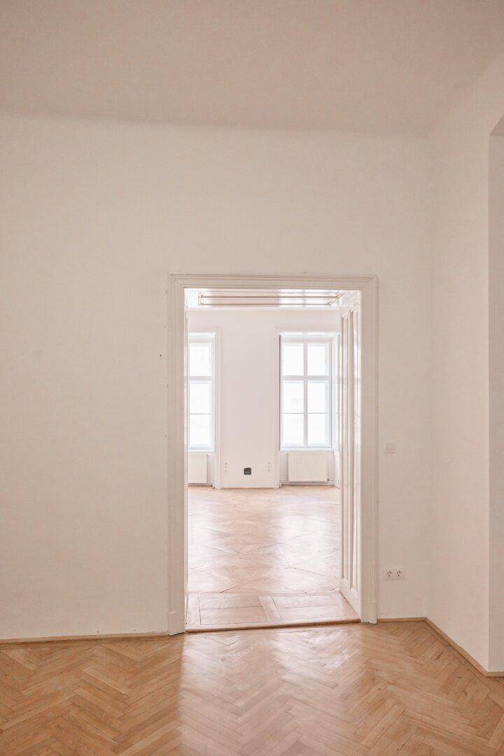 Heller Raum mit offenem Durchgang zu einem weiteren Zimmer mit Fenstern.