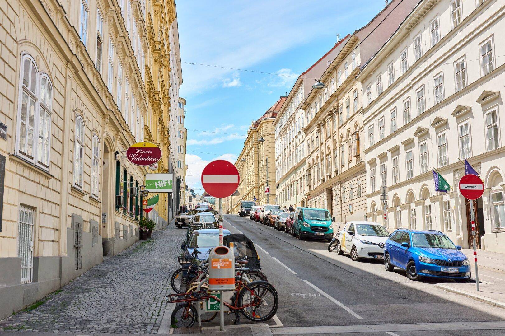Belebte Straße in einer europäischen Stadt mit historischen Gebäuden und geparkten Autos.