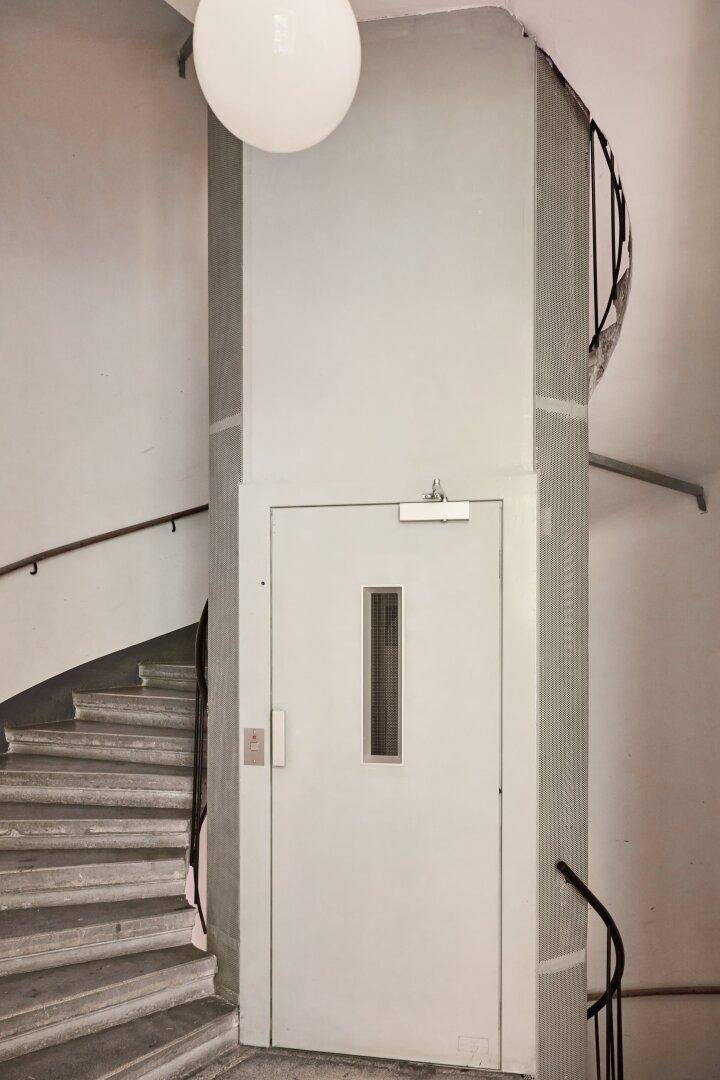 Geschwungene Steintreppe in einem Altbau mit einem modernen Aufzug im Treppenhaus.