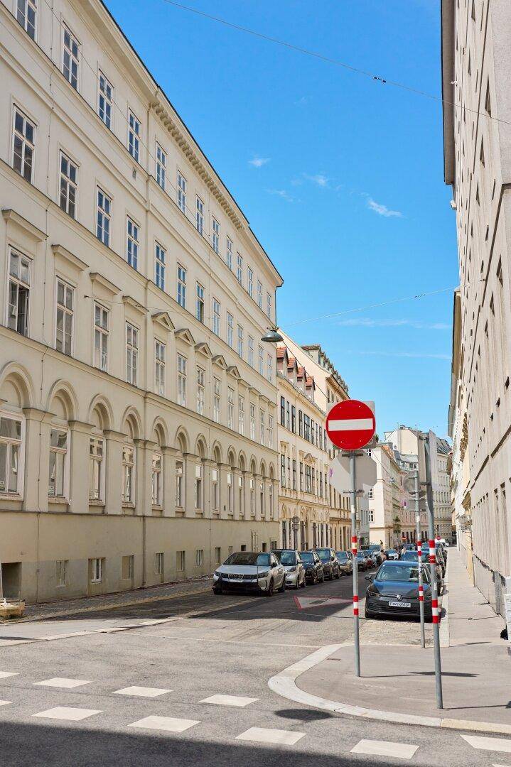 Städtische Straße mit historischen Gebäuden, geparkten Fahrzeugen und blauem Himmel.