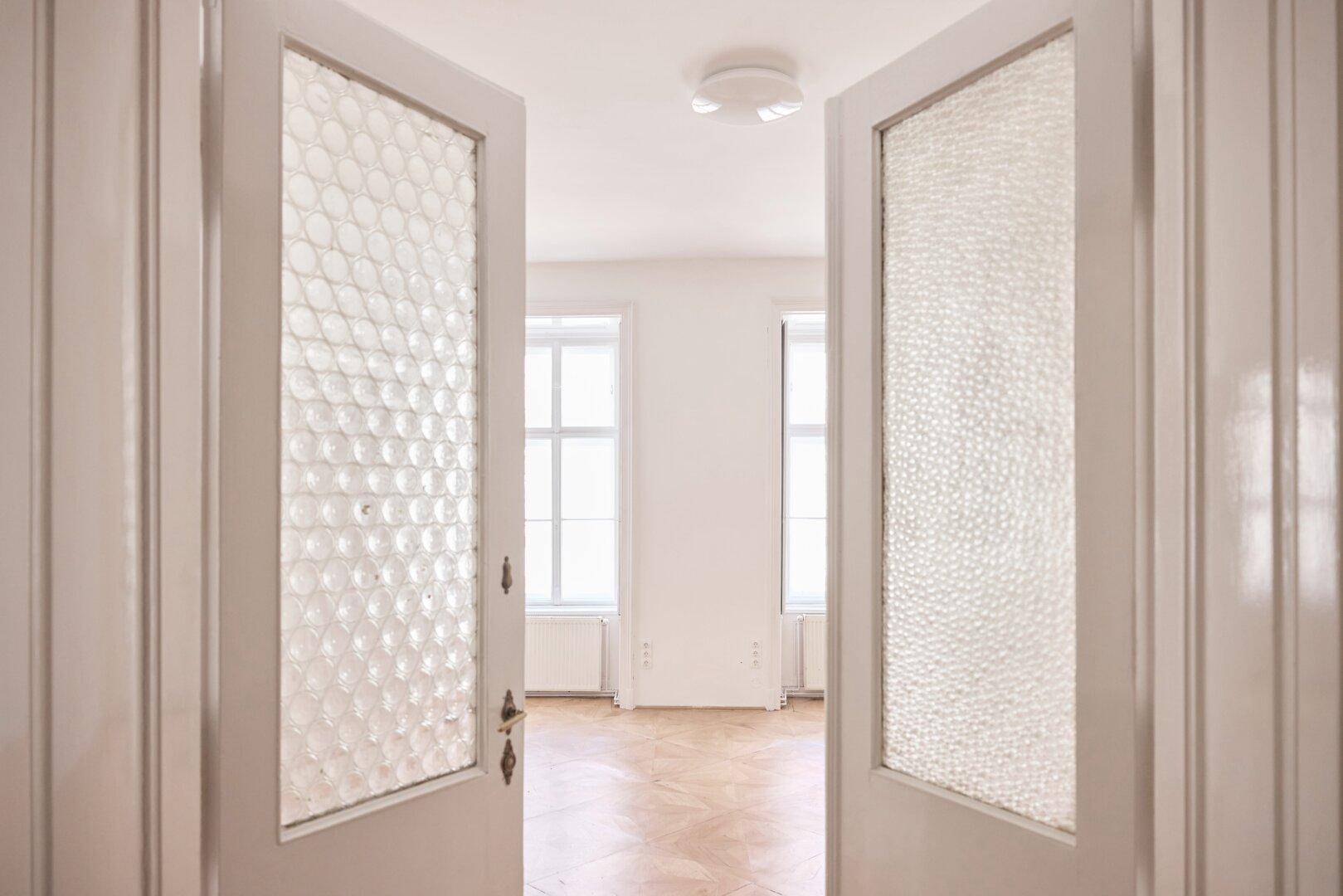 Blick durch eine Doppeltür mit strukturiertem Glas in einen hellen Raum mit Parkettboden und Fenstern.