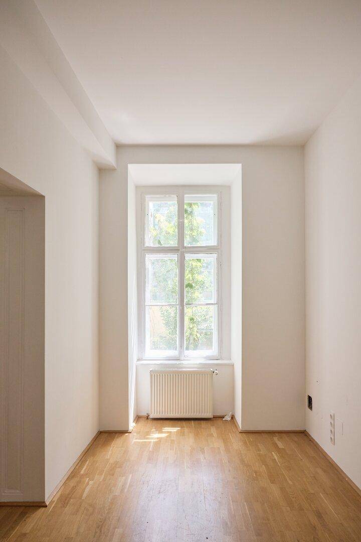 Heller, leerer Raum mit hohen Decken, Fischgrätparkett und großem Fenster mit Blick ins Grüne.