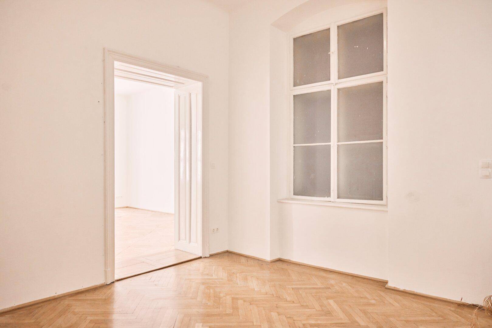 Heller, leerer Raum mit Fischgrätparkett und großem Fenster, Blick in einen weiteren Raum.
