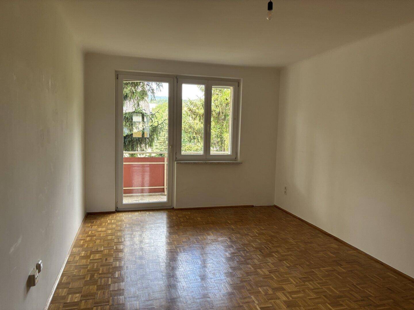 Helles Zimmer mit Parkettboden und Zugang zu einem Balkon, umgeben von grünen Bäumen.