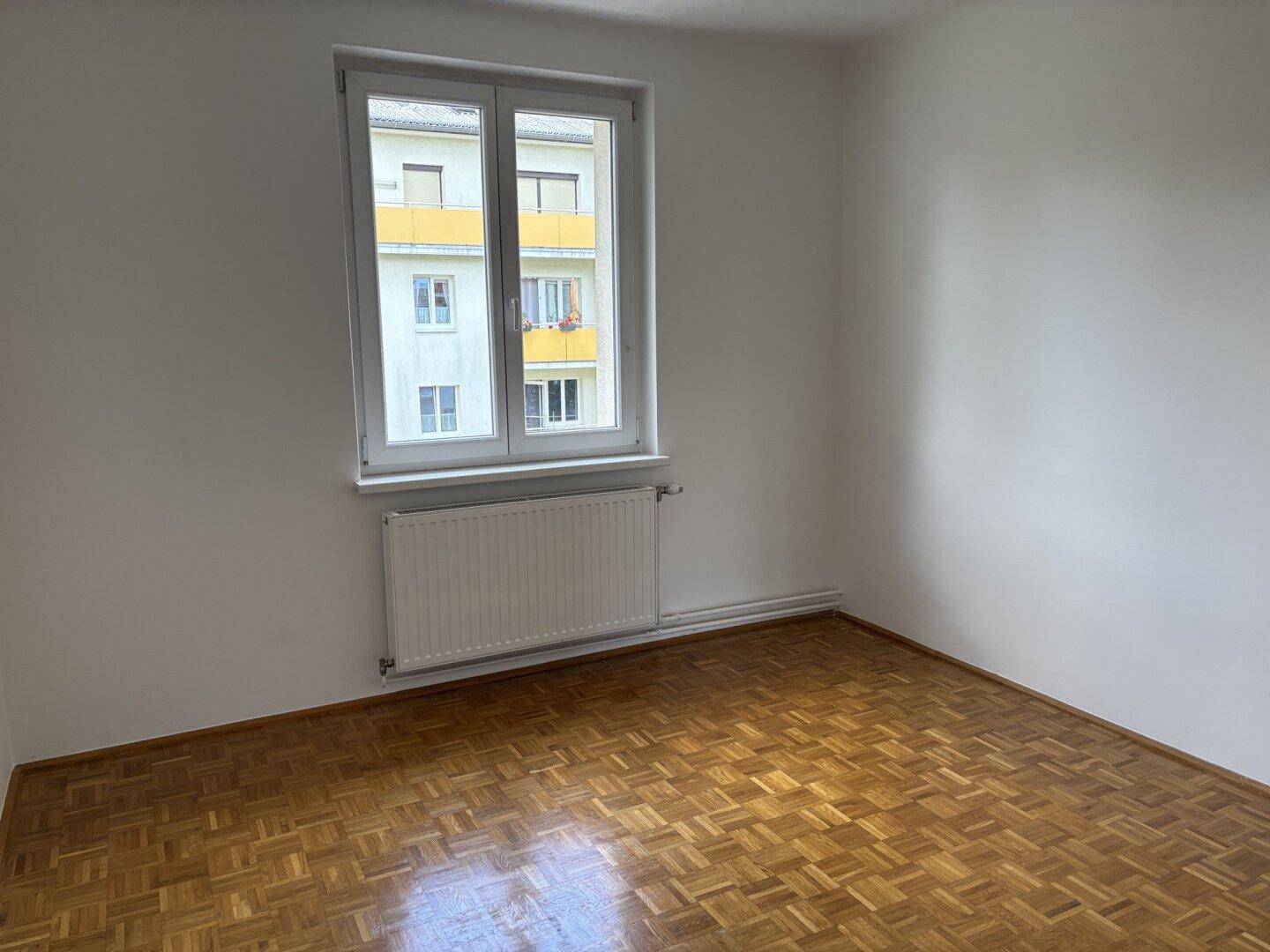 Leeres Zimmer mit Parkettboden, weiß gestrichenen Wänden und einem Fenster mit Blick auf ein Gebäude.