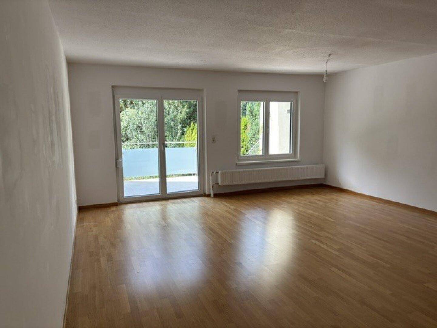 Geräumiges Wohnzimmer mit hellem Parkettboden, großen Fenstern und Zugang zum Balkon.