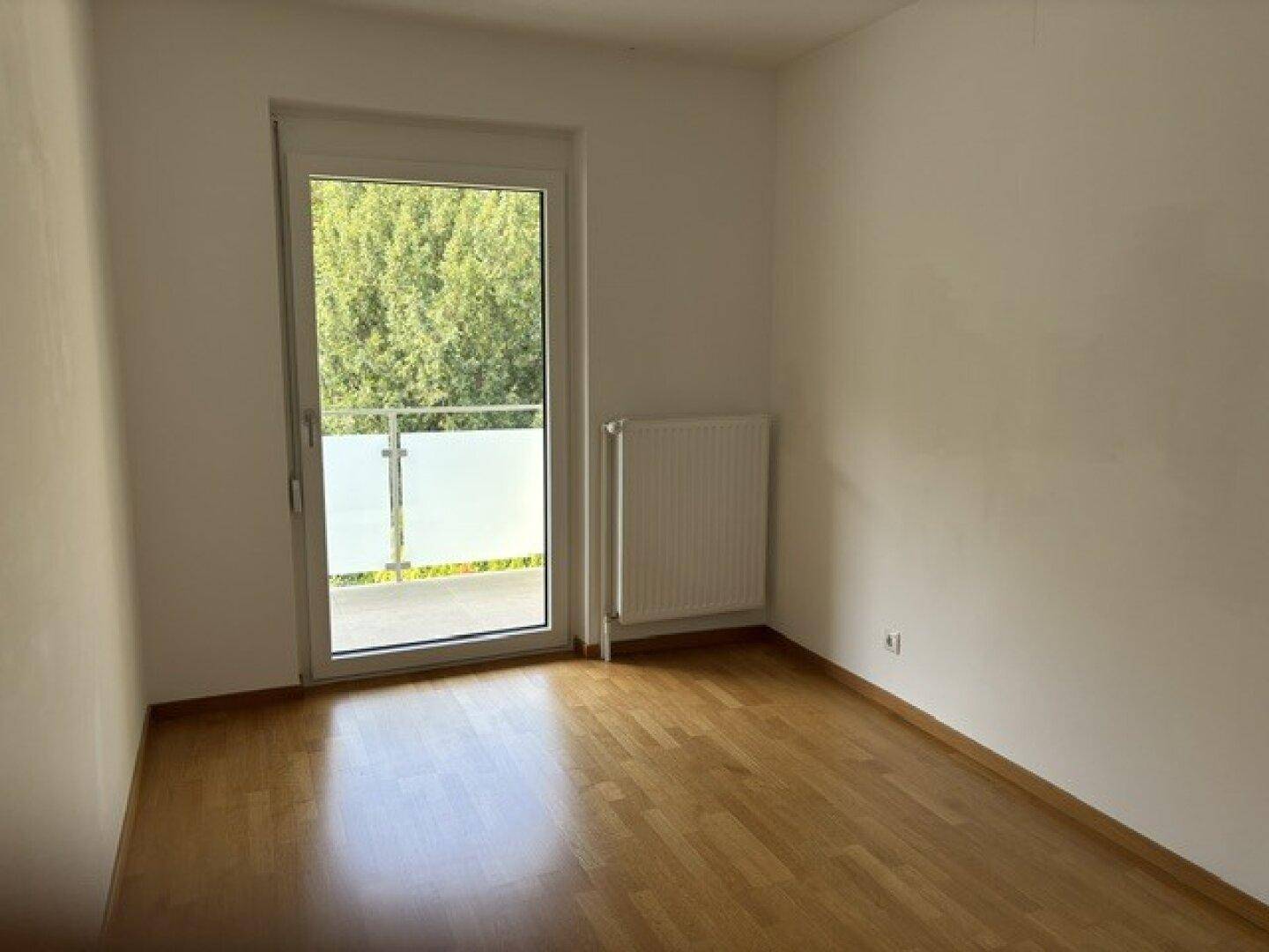 Helles Schlafzimmer mit Holzboden und Balkontür, die einen Blick ins Grüne bietet.