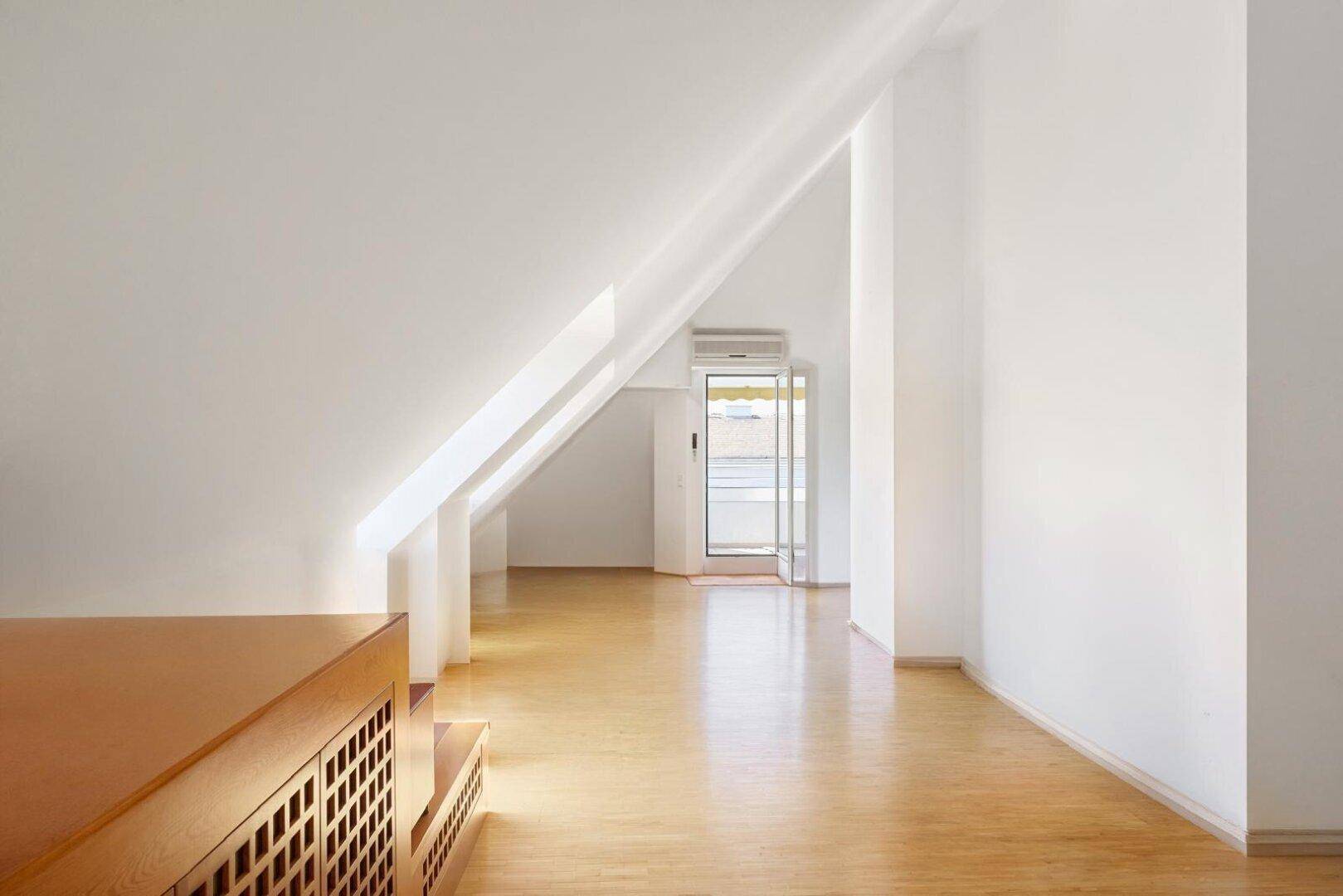 Heller Raum im Dachgeschoss mit schrägen Wänden, Holzboden und Zugang zum Balkon.
