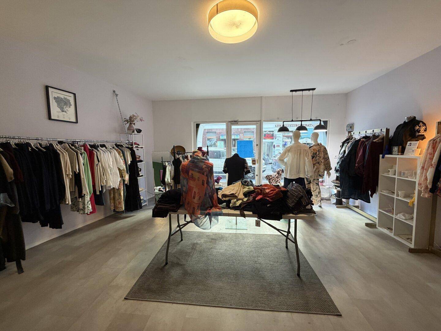 Heller Verkaufsraum mit Kleiderständern, Schaufenster und einem zentralen Präsentationstisch.
