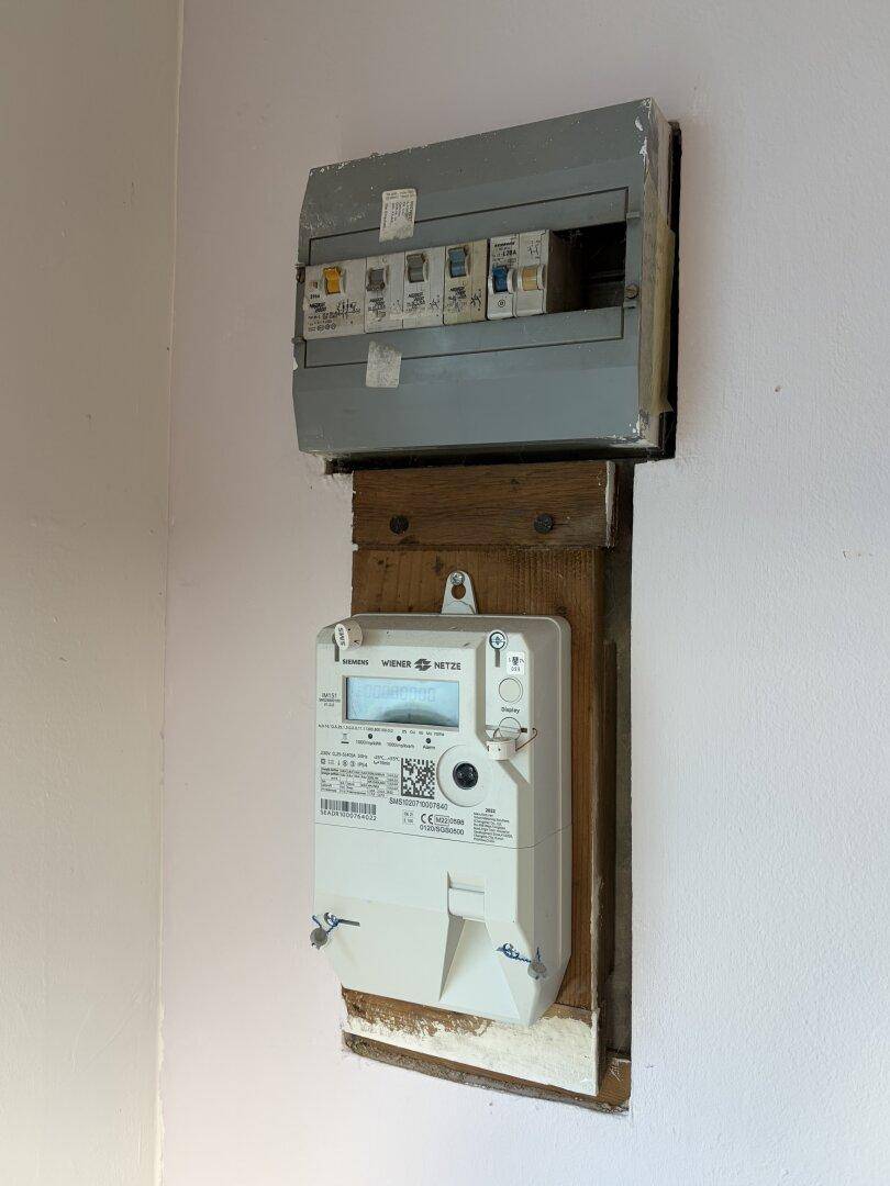 Elektrischer Zähler und Sicherungskasten an der Wand montiert, auf einer Holzplatte befestigt.