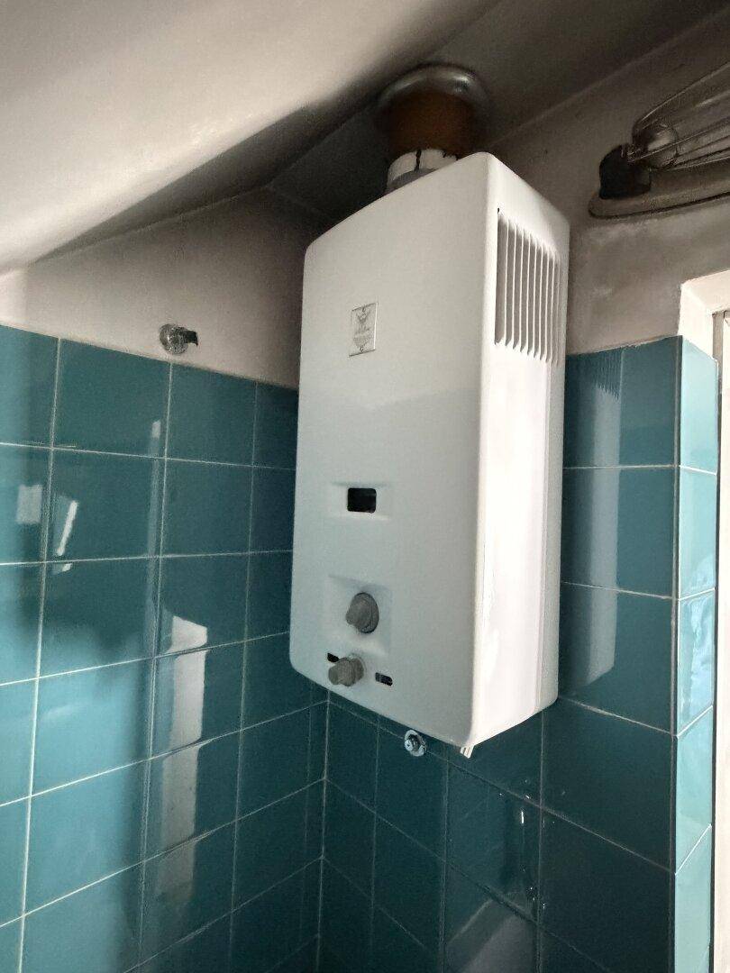 Wandmontierter Warmwasserbereiter in einem Raum mit alten, türkisfarbenen Fliesen.