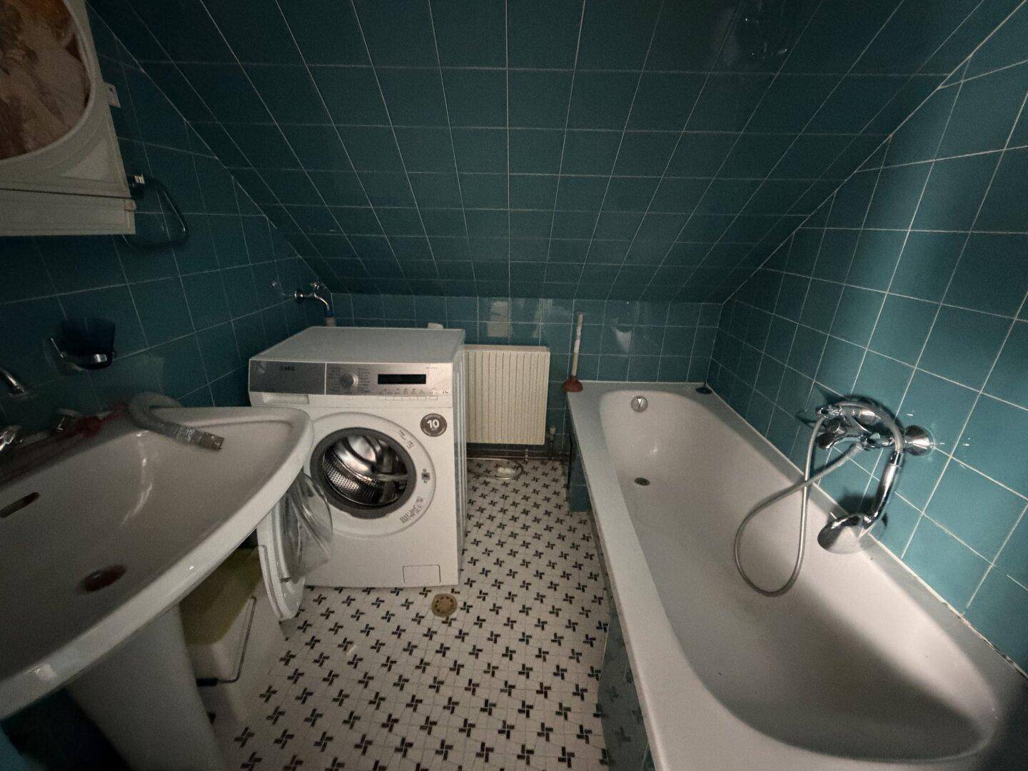 Badezimmer mit Badewanne, Waschmaschine und türkisfarbenen Wandfliesen unter einer Dachschräge.