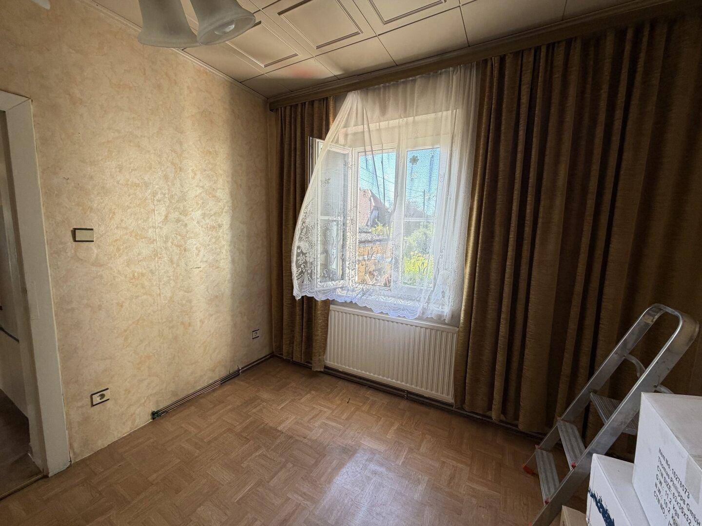 Schlichtes Zimmer mit gemusterter Tapete, Parkettboden und einem Fenster mit braunen Vorhängen.