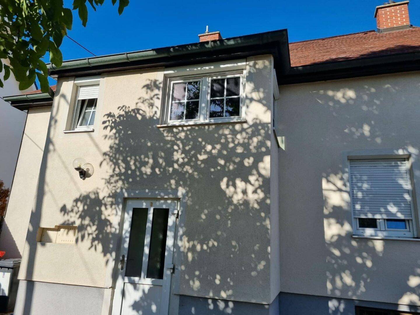 Rückseite des Hauses mit Fenstern und einer Tür, teilweise im Schatten von Bäumen.