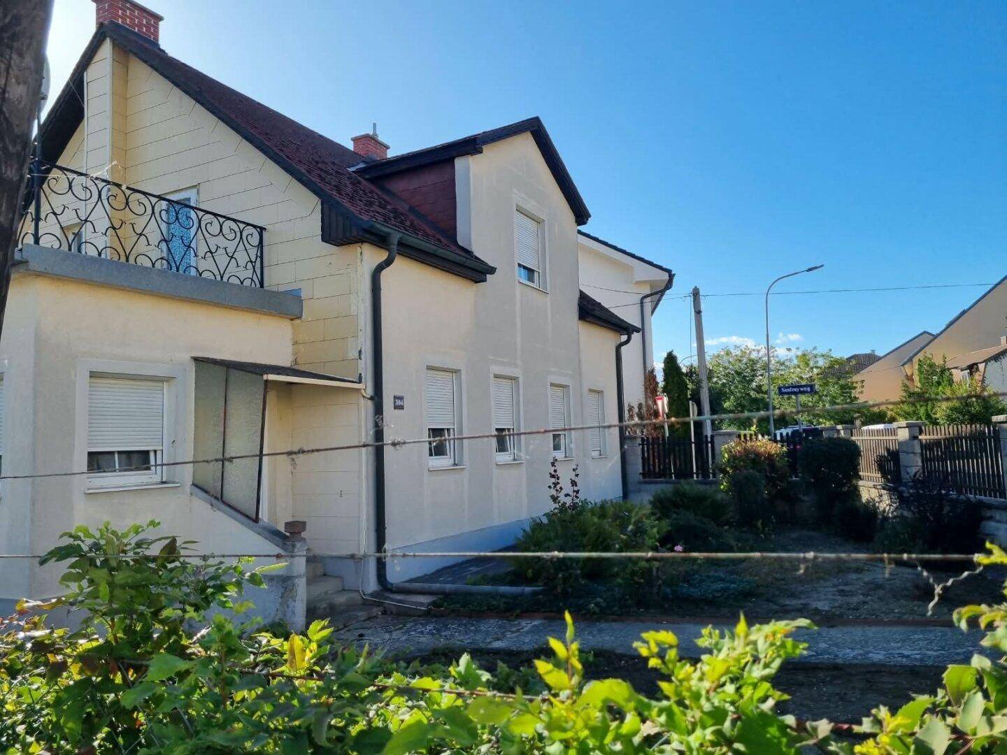 Seitenansicht eines Hauses mit Balkon und Rollläden, umgeben von einem Zaun und Bäumen.
