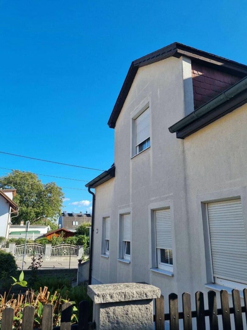 Seitenfassade des Hauses mit mehreren Fenstern und geschlossenen Rollläden, umgeben von einem Zaun.