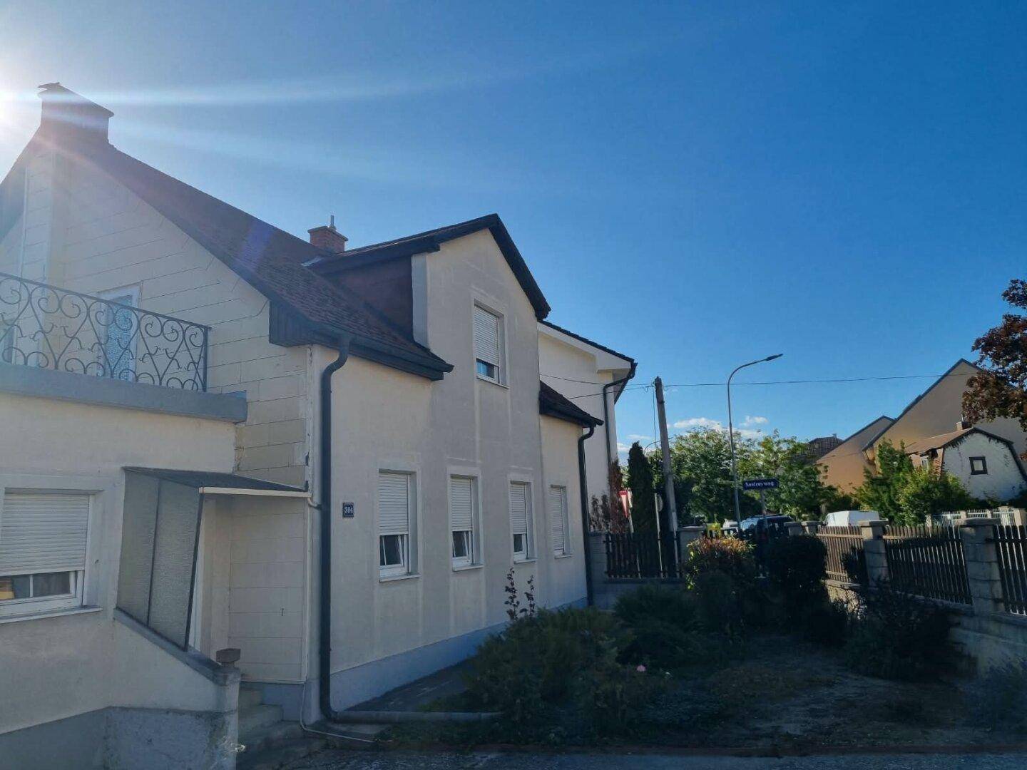 Seitenansicht des Hauses mit Rollläden und einem Balkon, umgeben von grüner Vegetation und einem Zaun.