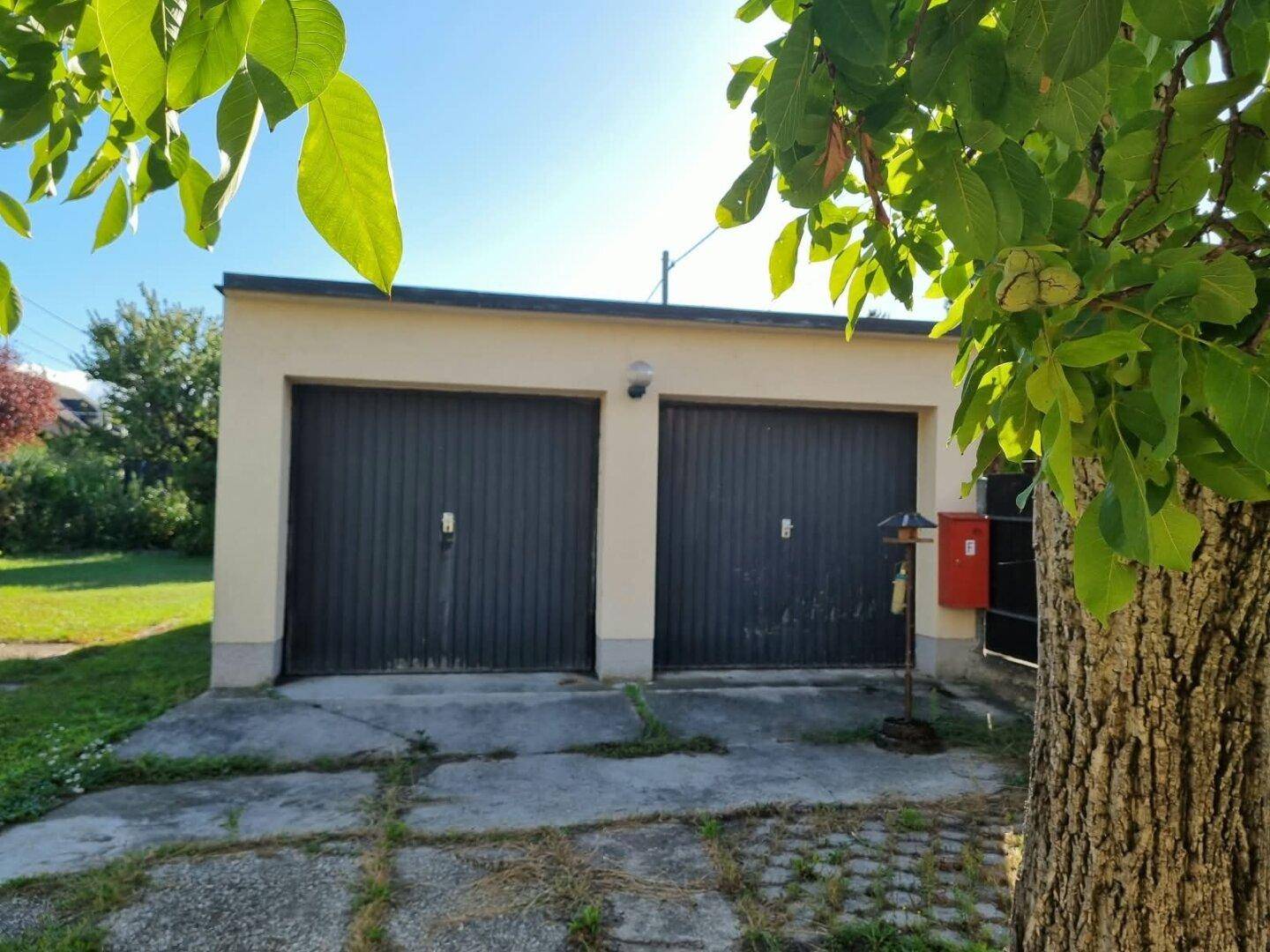 Doppelgarage mit zwei dunklen Toren und einem gepflasterten Vorplatz unter hellem Sonnenlicht.