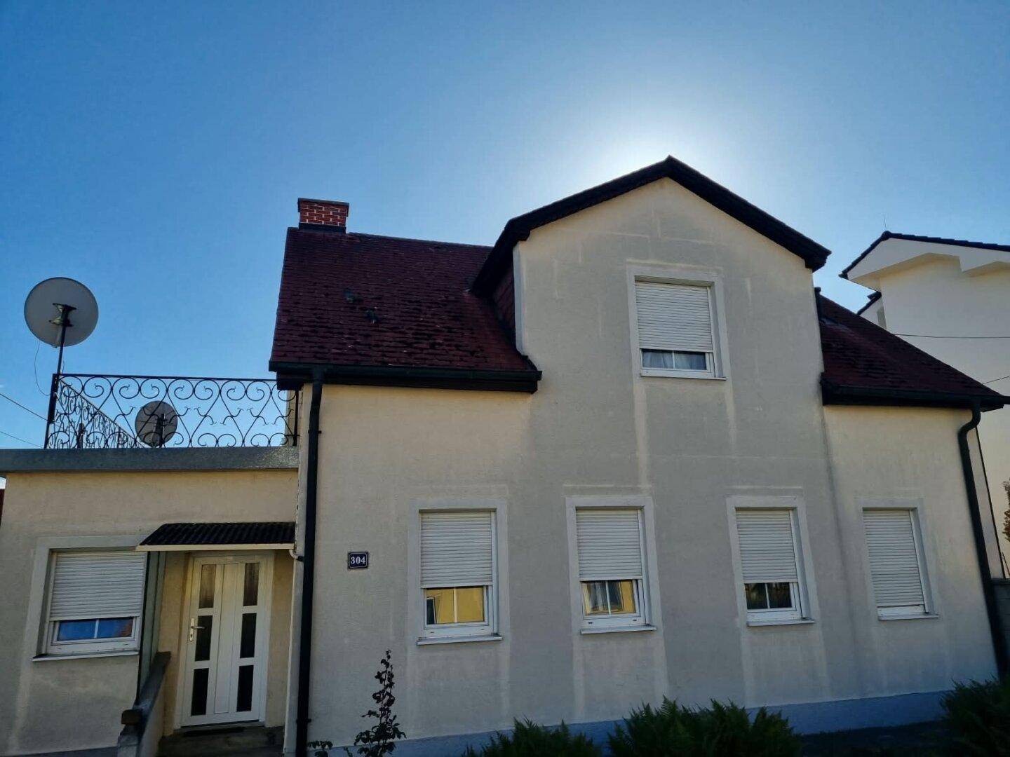 Vorderansicht des Hauses mit Eingangsbereich, Balkon und geschlossenen Rollläden unter strahlendem Himmel.