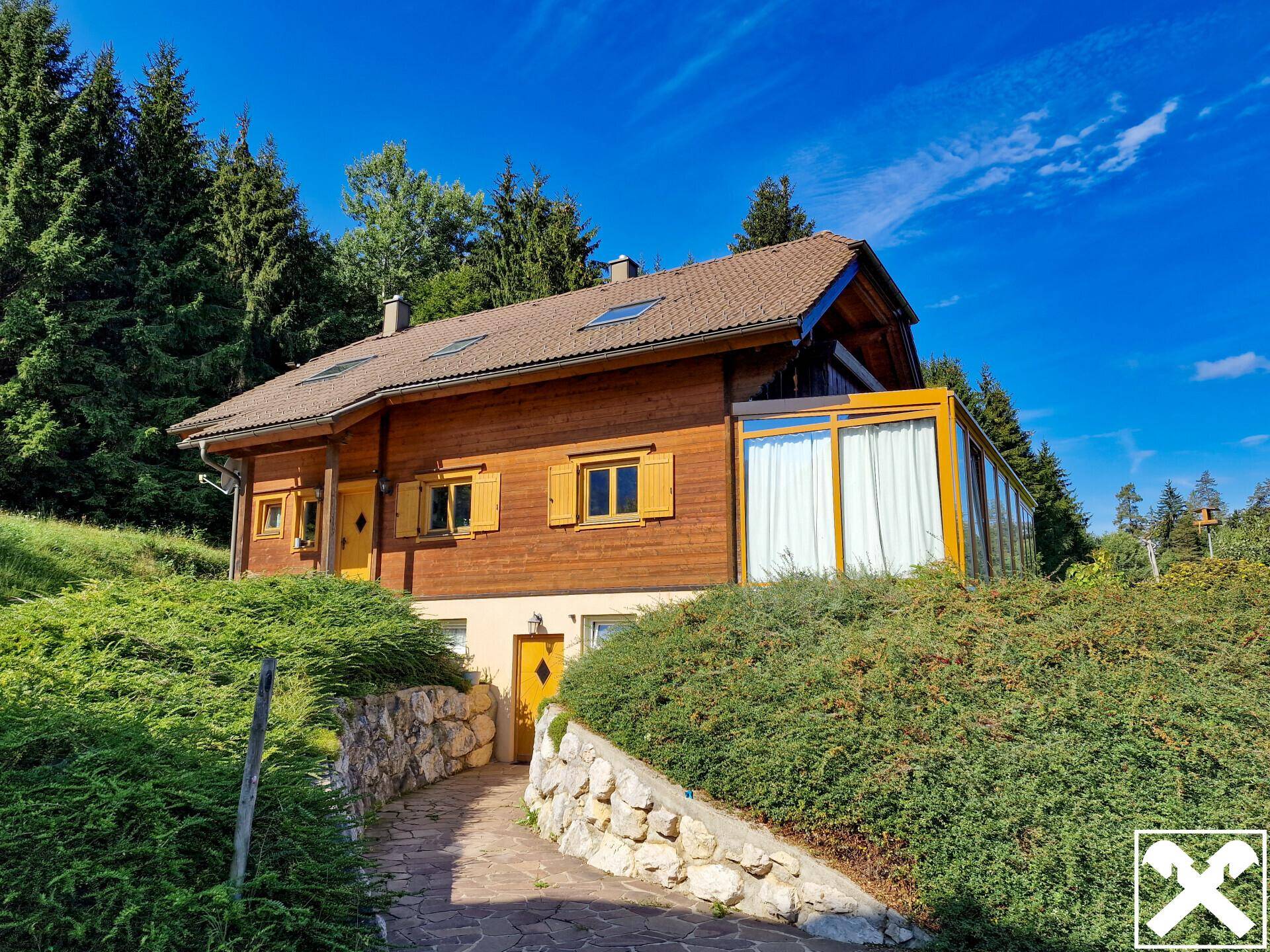 Holzhaus mit sonnigem Wintergarten und gepflegter Auffahrt, umgeben von grünen Büschen.