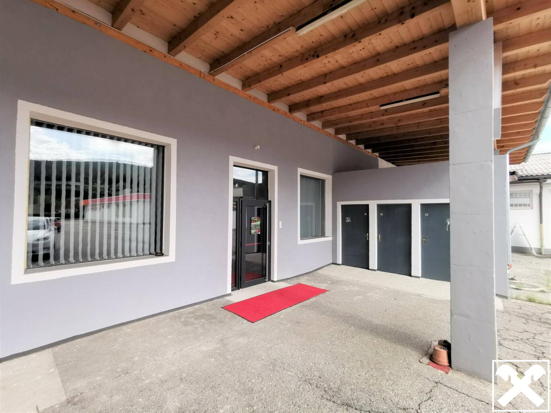 Eingangsbereich eines Gebäudes mit überdachtem Zugang, grauer Fassade, Holzbalkendecke und mehreren Türen.