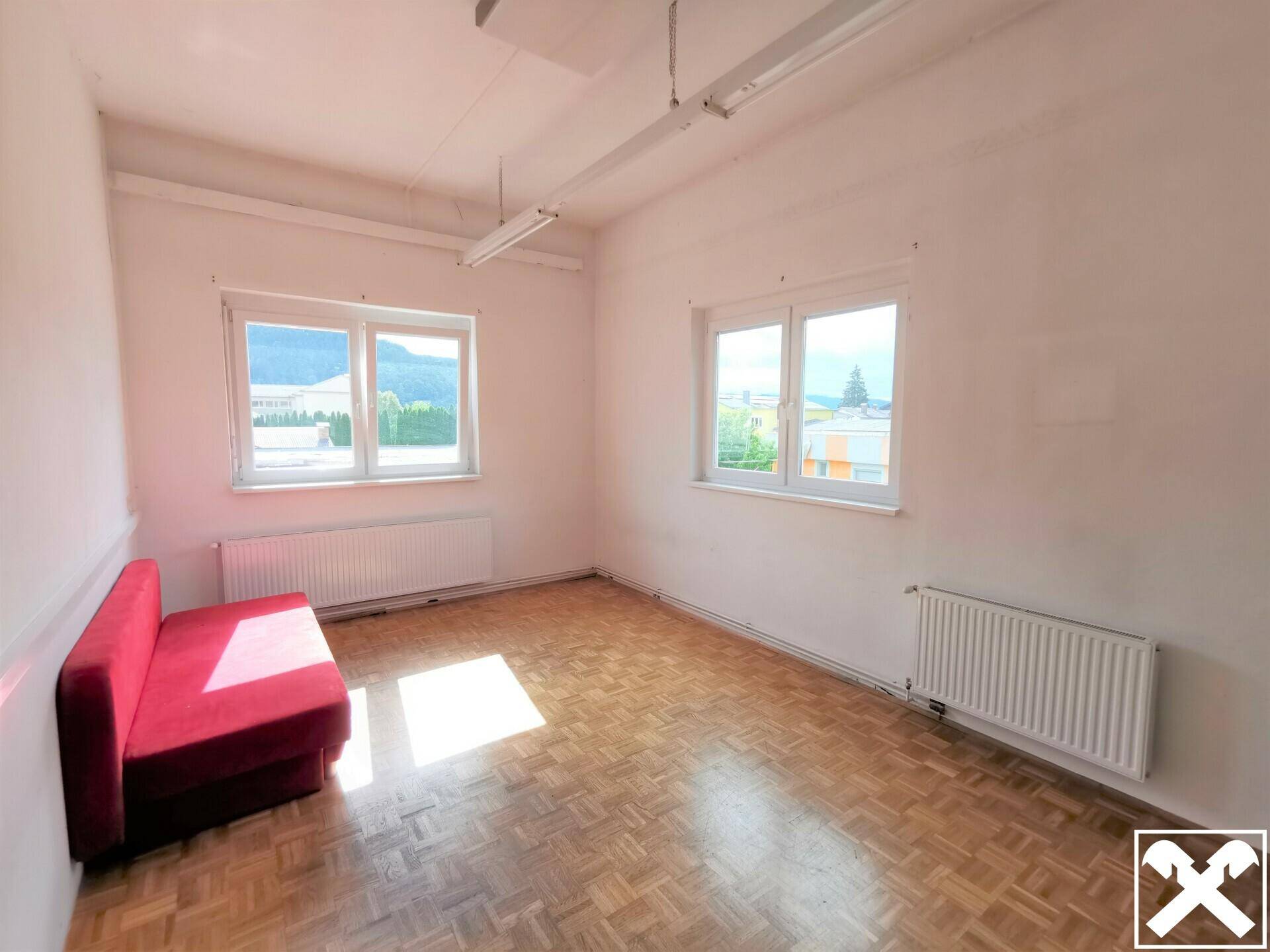 Heller Raum mit Holzboden, zwei großen Fenstern und einem roten Sofa, ideal als Büro oder Aufenthaltsraum.