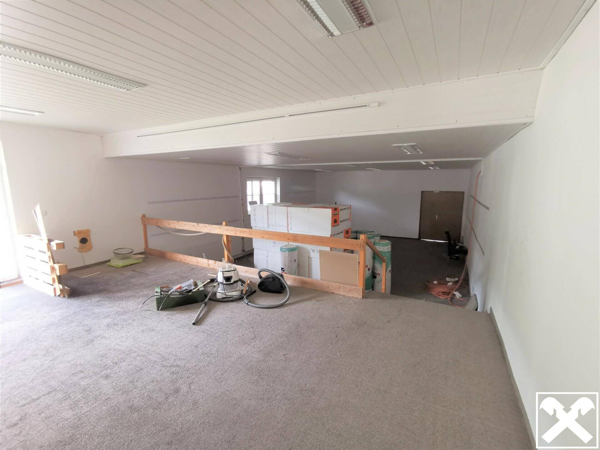 Großer, offener Gewerberaum mit Teppichboden und einer abgesenkten Fläche im Hintergrund, ideal für Lagerung oder Werkstatt.