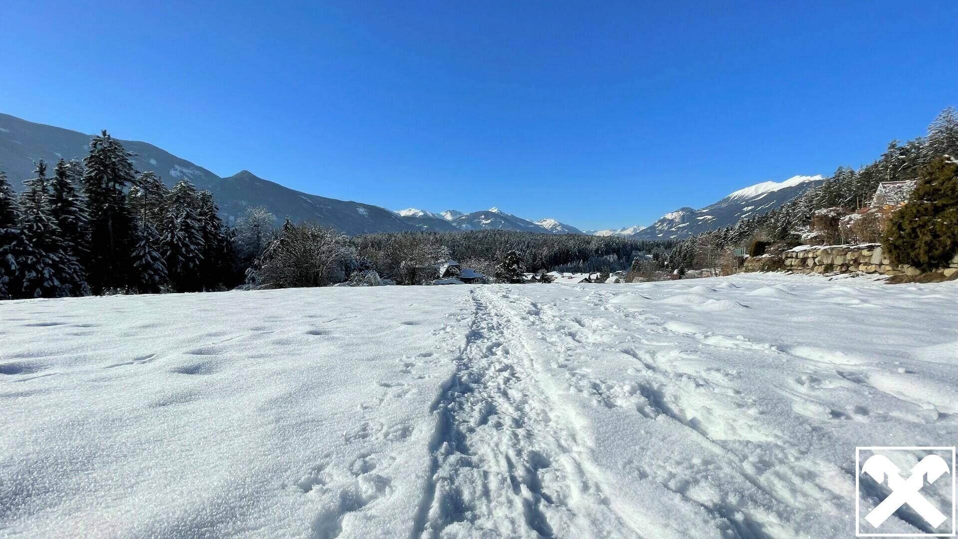 Verschneite Winterlandschaft mit Spuren im Schnee, umgeben von Wäldern und schneebedeckten Bergen.