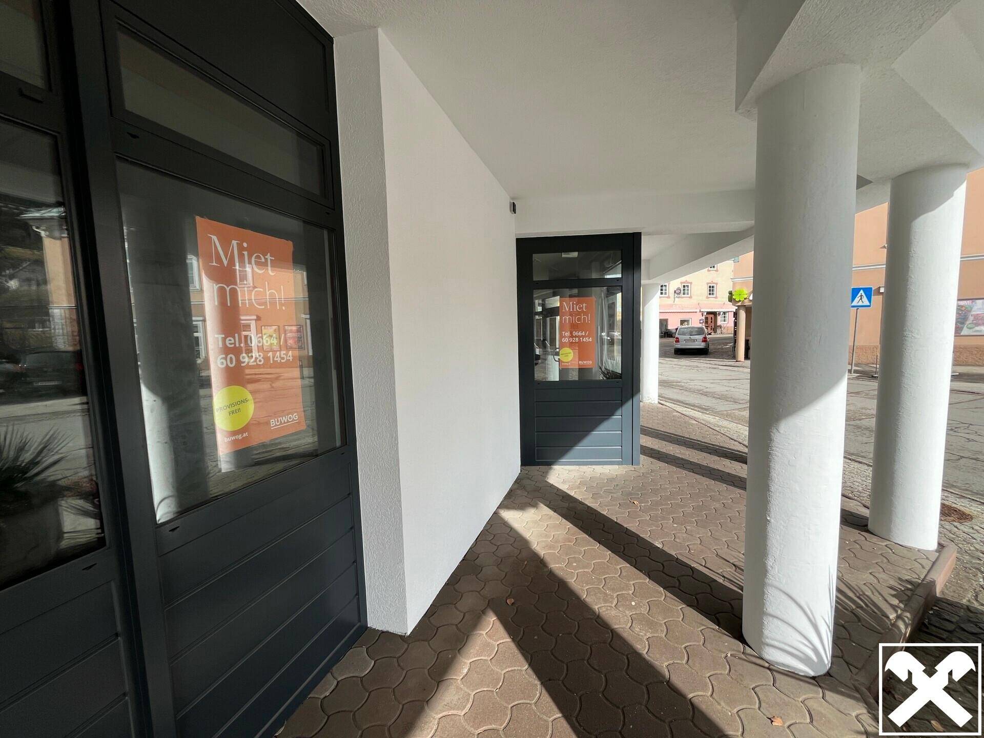 Überdachter Eingangsbereich eines Geschäftsgebäudes mit Säulen und großen Fenstern, die zur Straße zeigen.