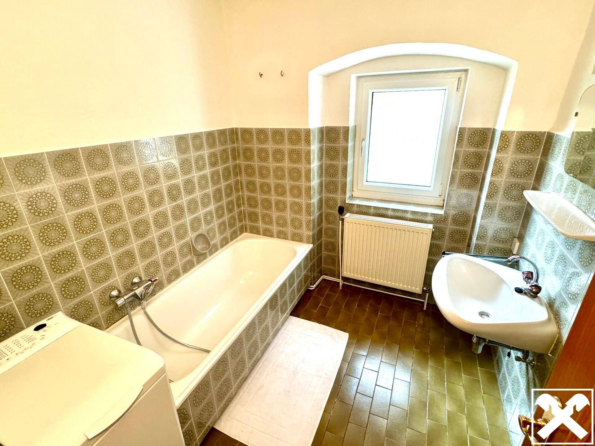 Badezimmer mit Badewanne, Waschbecken, Fenster und älteren Fliesen im Retro-Stil der 80er Jahre.