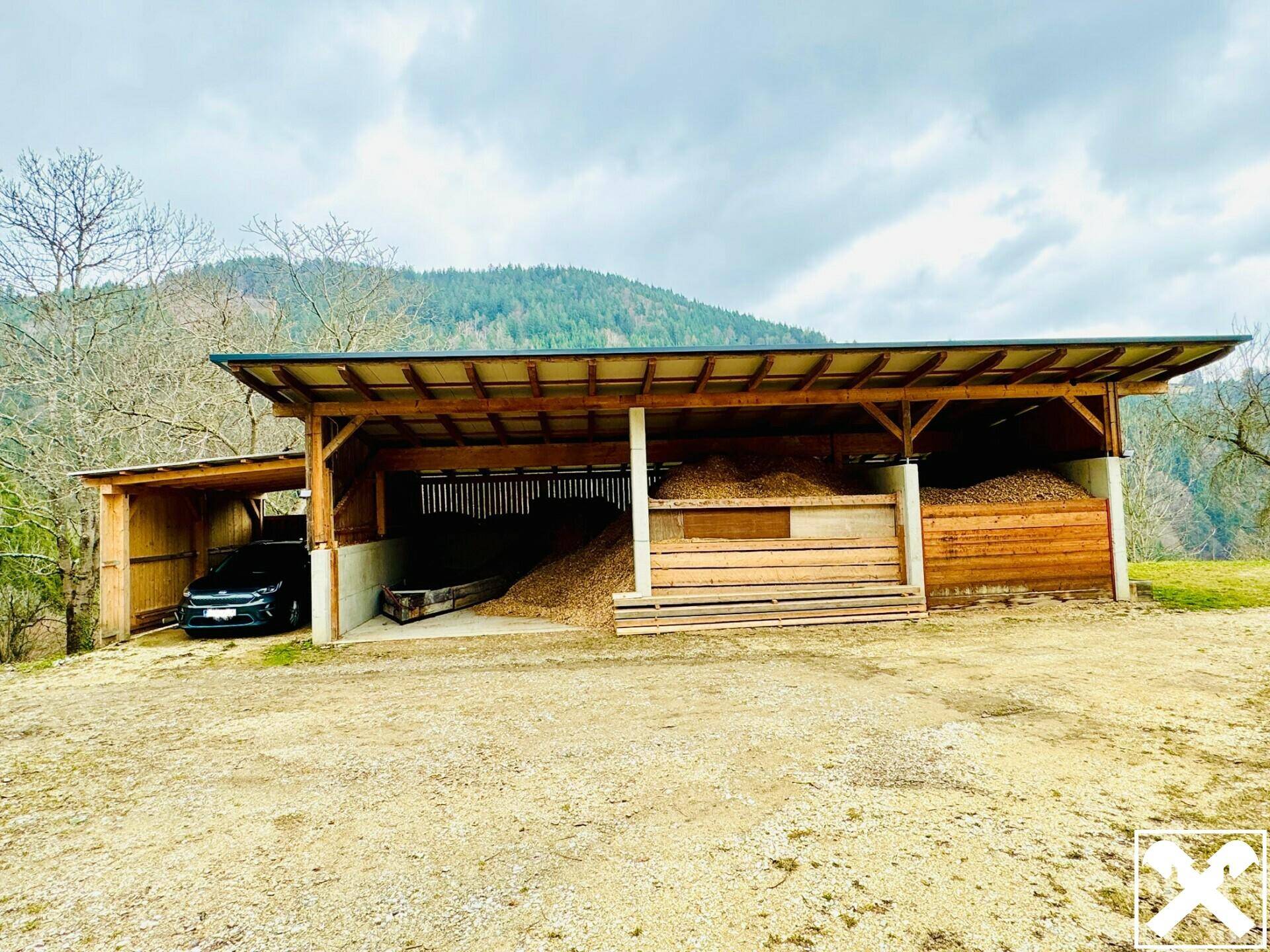 Offener Holzschuppen mit gelagerten Holzspänen und einem geparkten Fahrzeug in ländlicher Umgebung.
