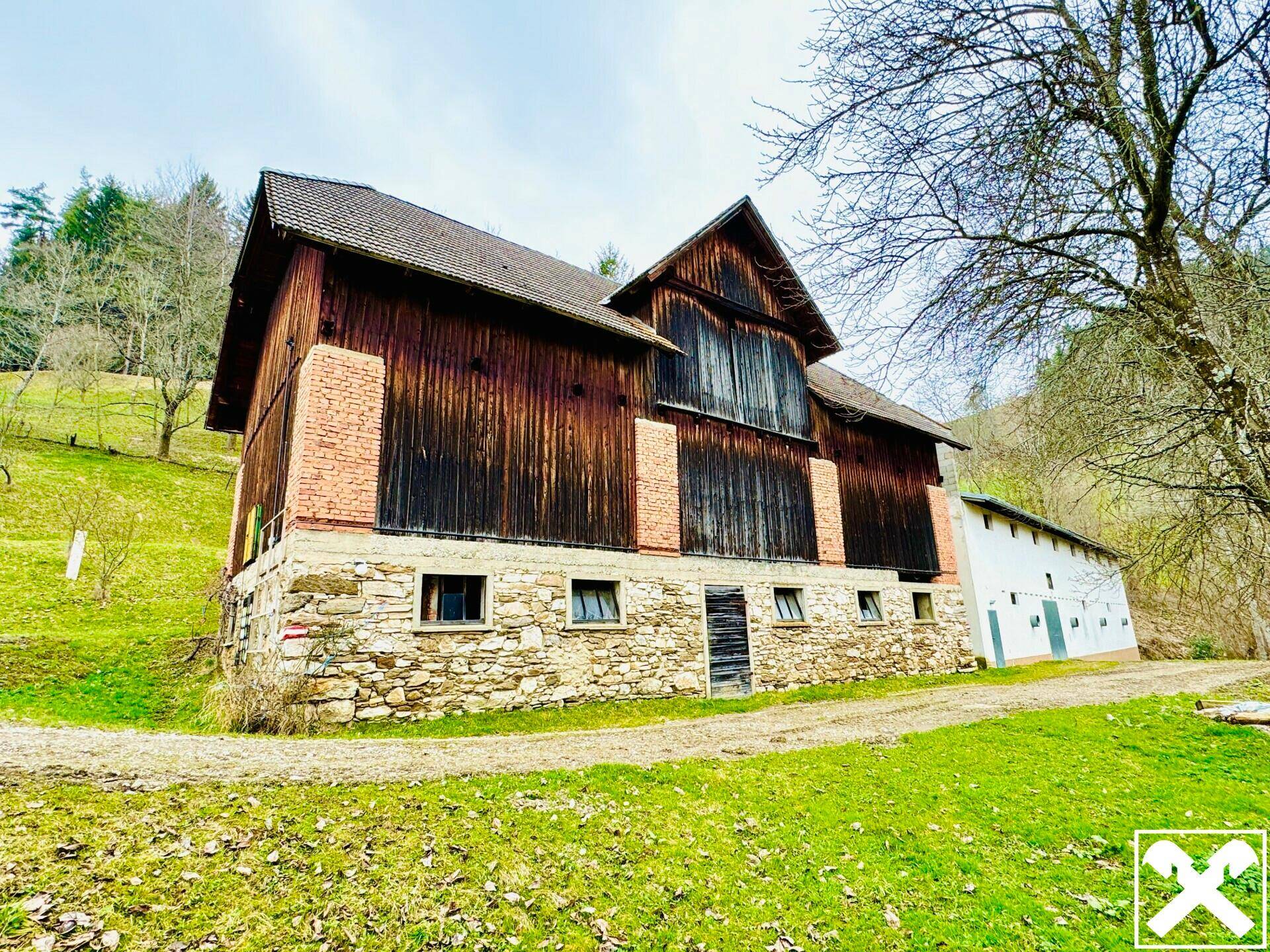 Rustikales Bauernhaus mit Holzwänden, Steinfundament und Ziegeldetails, eingebettet in eine grüne Landschaft.