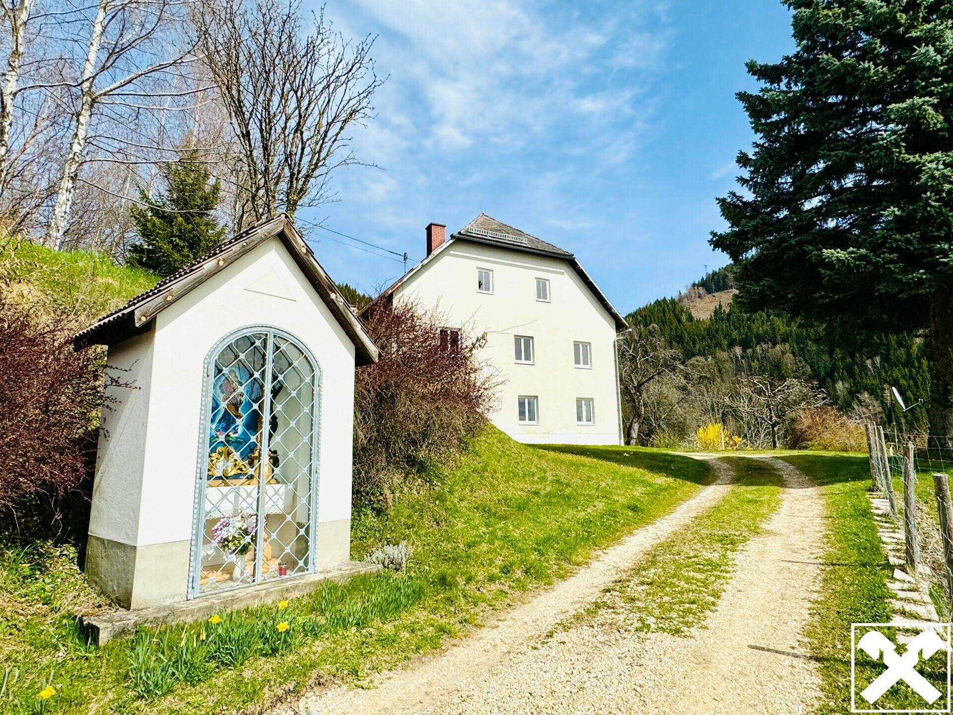 Ländliches Wohnhaus mit kleinem Wegschrein und grüner Umgebung unter strahlendem Himmel.