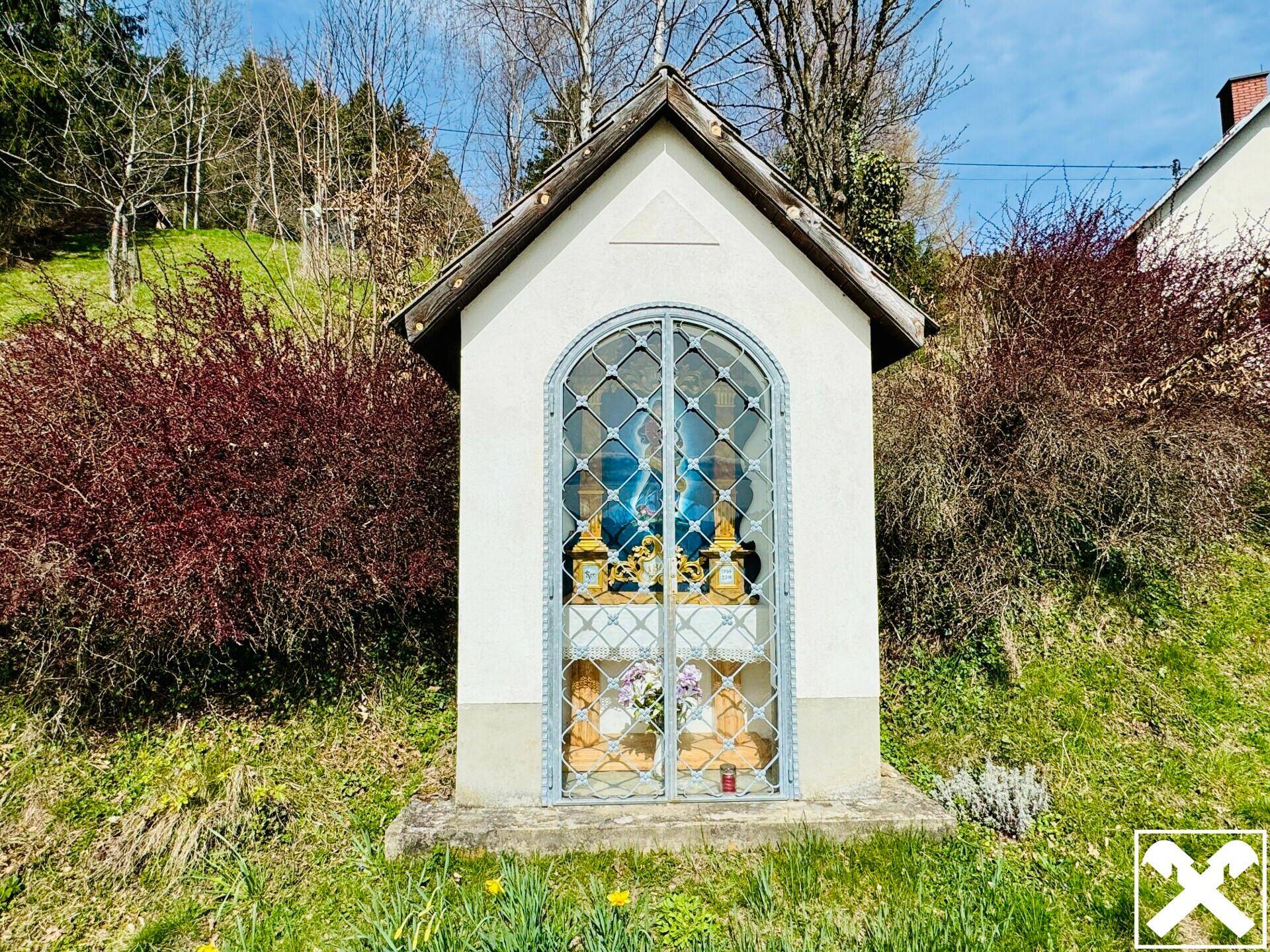 Kleiner weißer Wegschrein mit Gittertür und religiöser Darstellung, eingebettet in eine grüne Hügellandschaft.