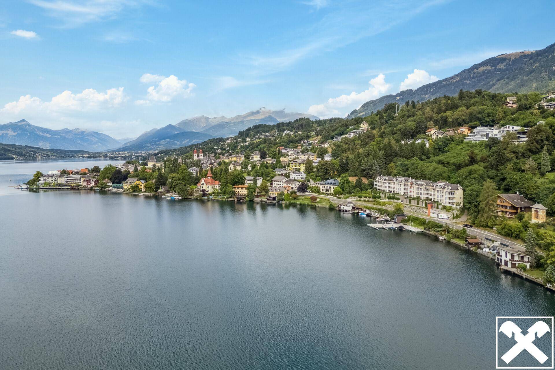 Panoramablick auf den See mit umliegenden Häusern und bewaldeten Bergen unter blauem Himmel.