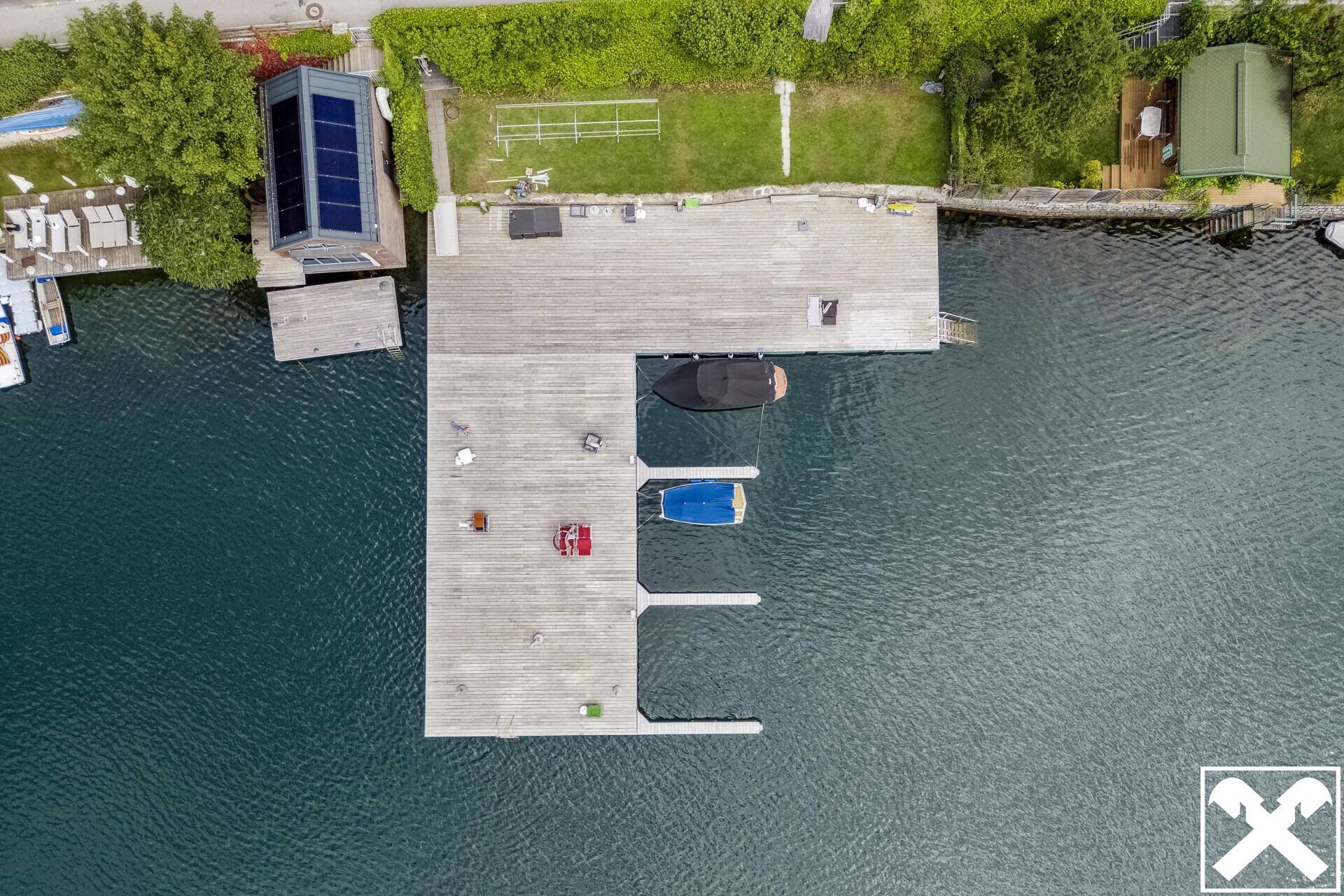 Luftaufnahme eines Holzstegs mit Bootsanlegestellen und angrenzender Grünfläche am Seeufer.