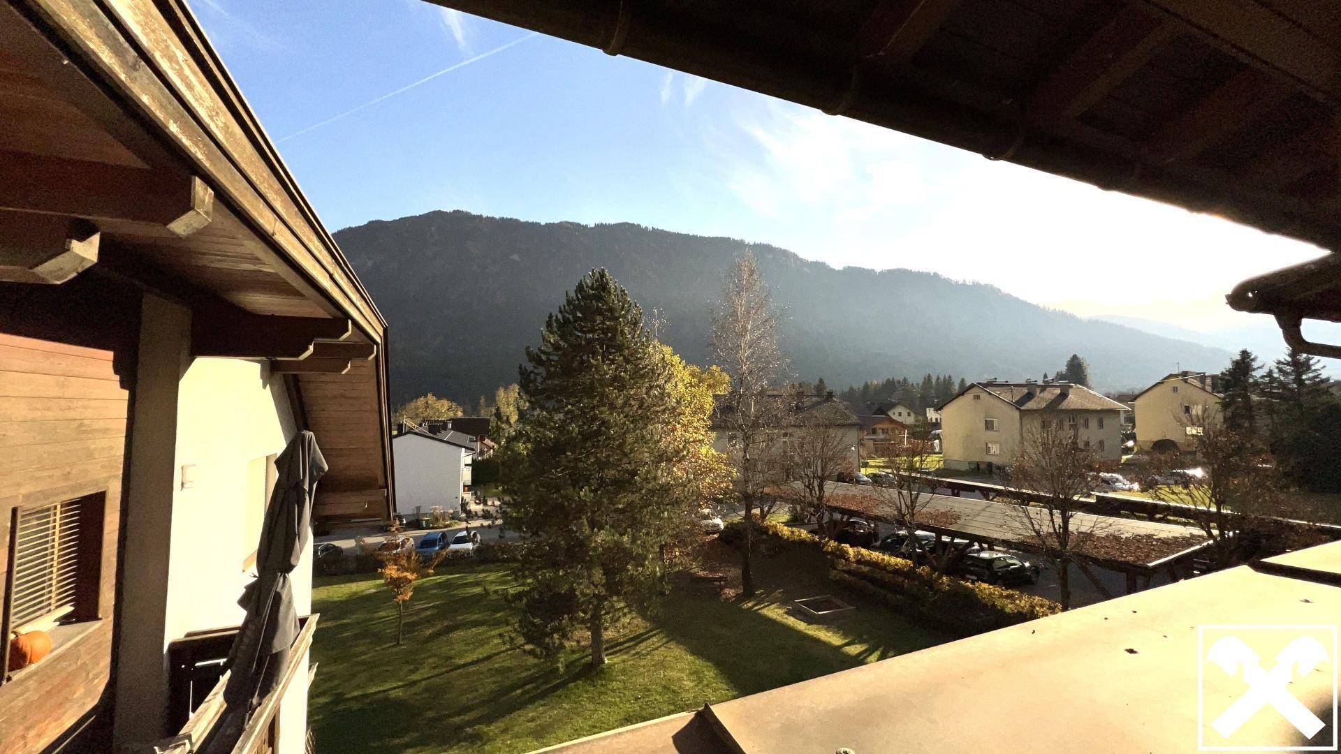 Sonniger Ausblick von einem Holzbalkon auf grüne Bäume und die dahinterliegende Bergkulisse am Morgen.