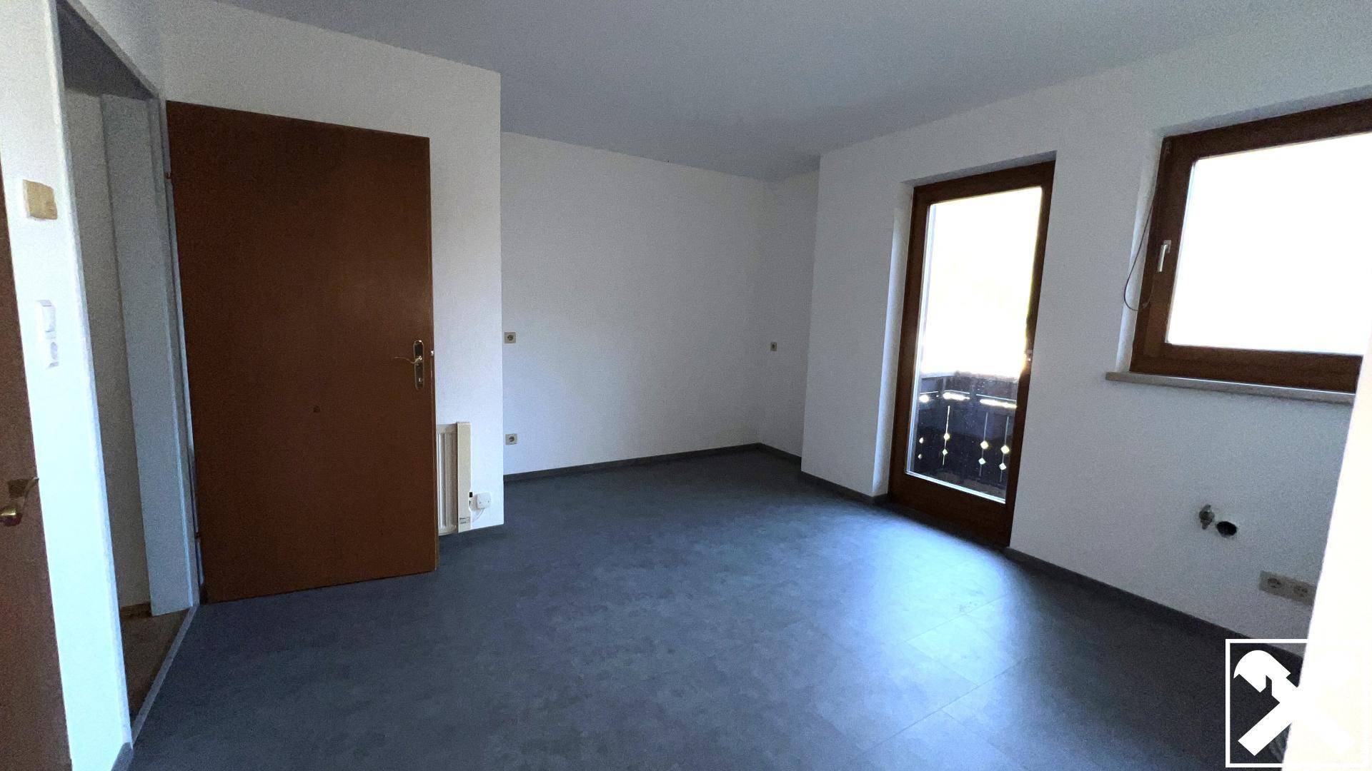 Geräumiges Zimmer mit dunklem Boden, weiß gestrichenen Wänden und Zugang zu einem Balkon oder einer Terrasse.