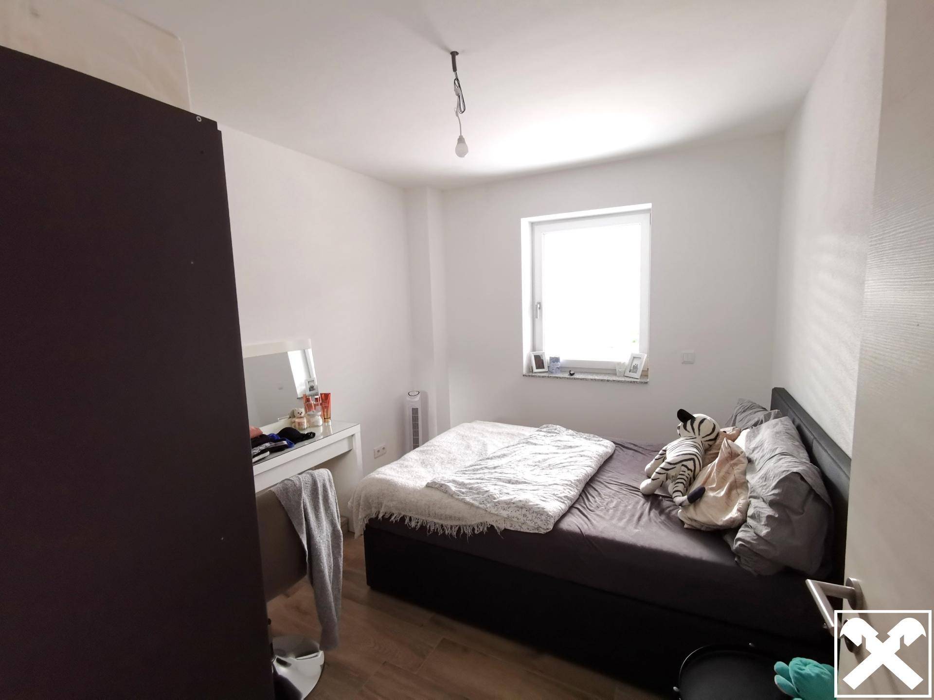 Helles Schlafzimmer mit Doppelbett, Schrank und Fenster, ausgestattet mit Holzoptikboden.