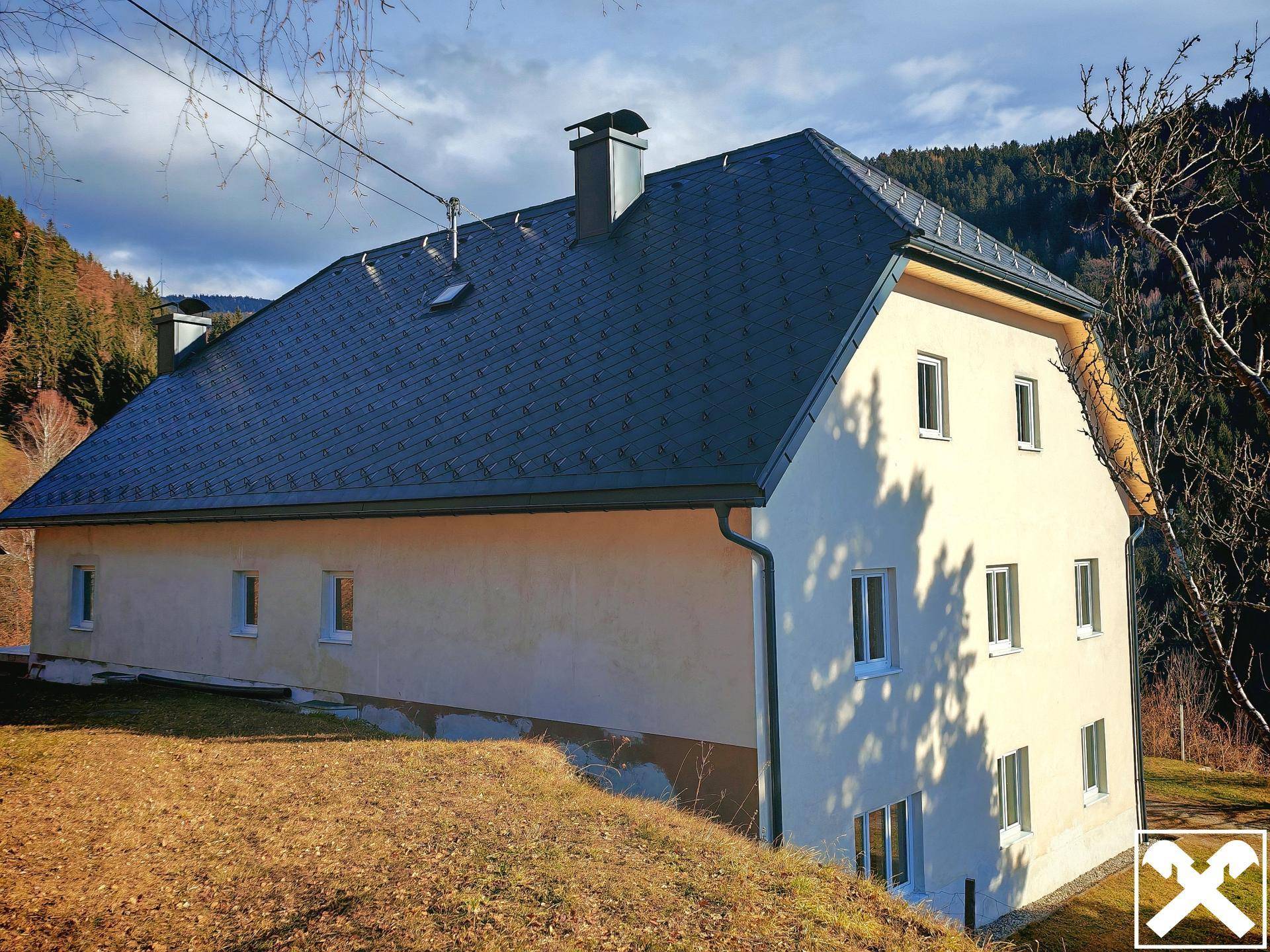 Haus mit dunklem Satteldach und heller Fassade in ländlicher Umgebung mit Bergblick.