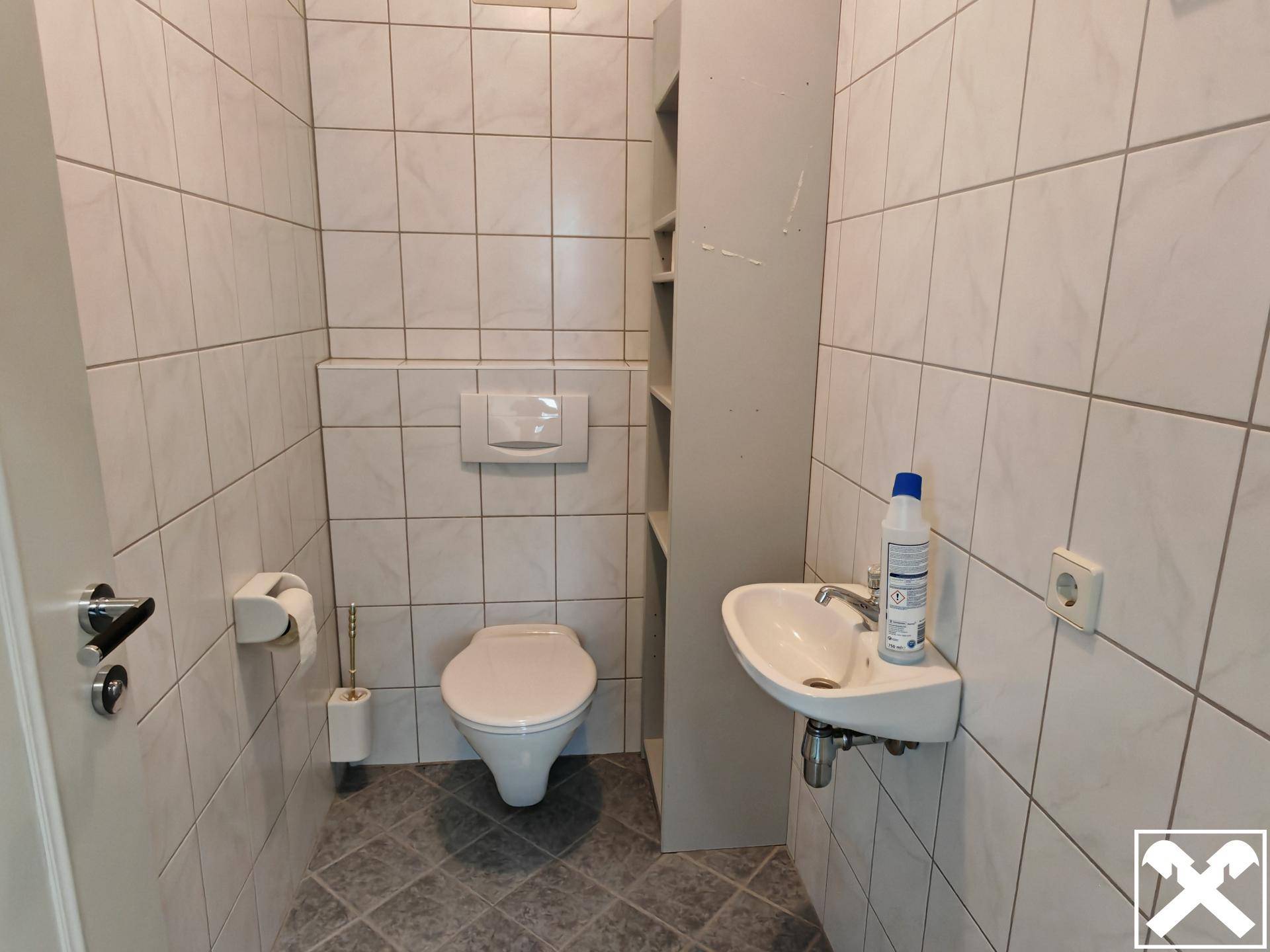 Gefliestes Gäste-WC mit wandhängender Toilette, Waschbecken und Ablageregal.