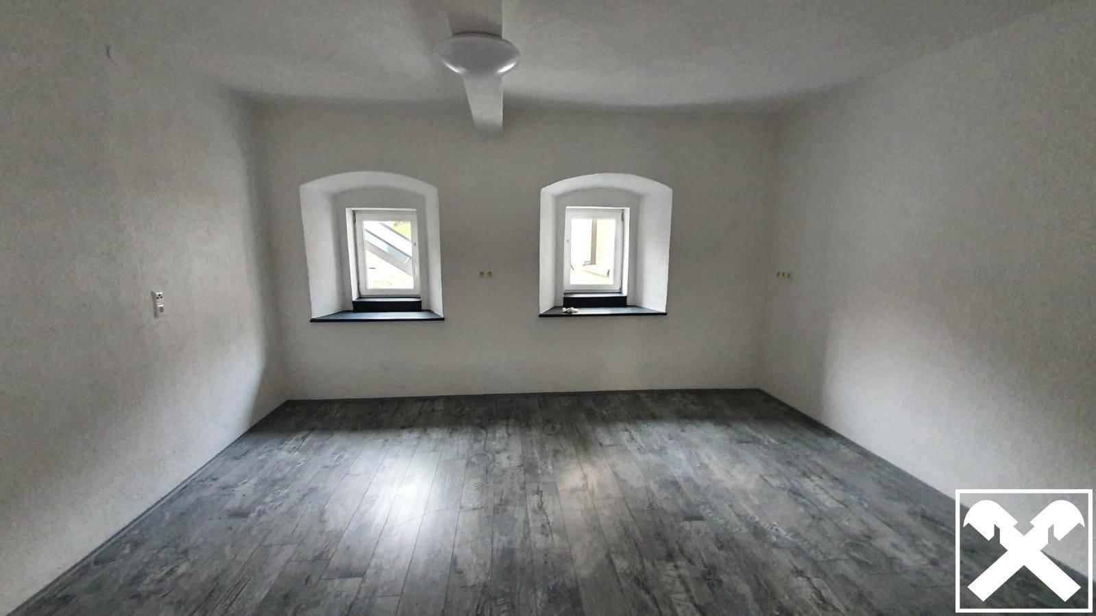 Leeres Zimmer mit zwei kleinen Fenstern und dunklem Laminatboden, ideal als Schlafzimmer oder Büro.