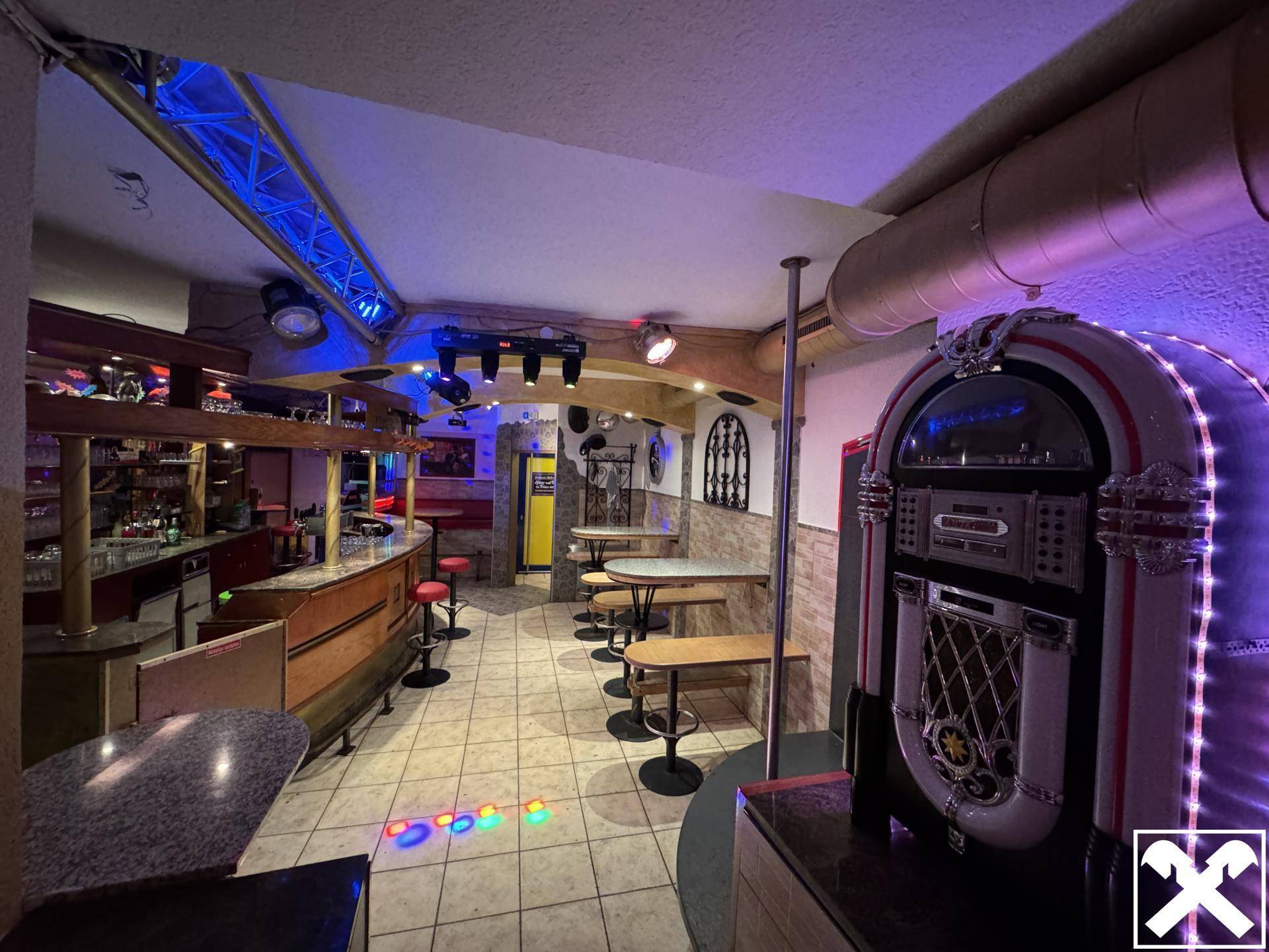 Innenansicht einer Bar mit Jukebox, Theke und Tanzfläche mit bunten Lichtern und Dekoration.