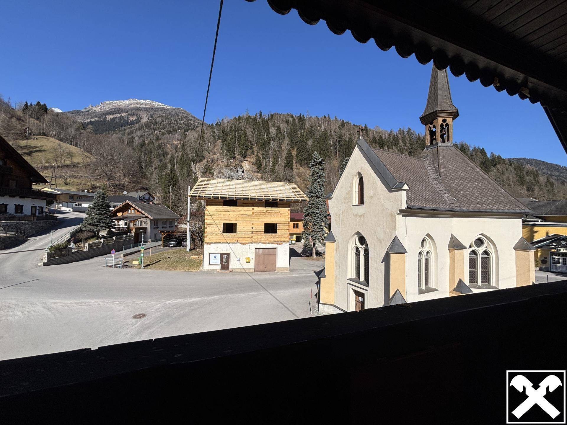 Blick vom Balkon auf eine kleine Kirche und die Berglandschaft im Sonnenschein.