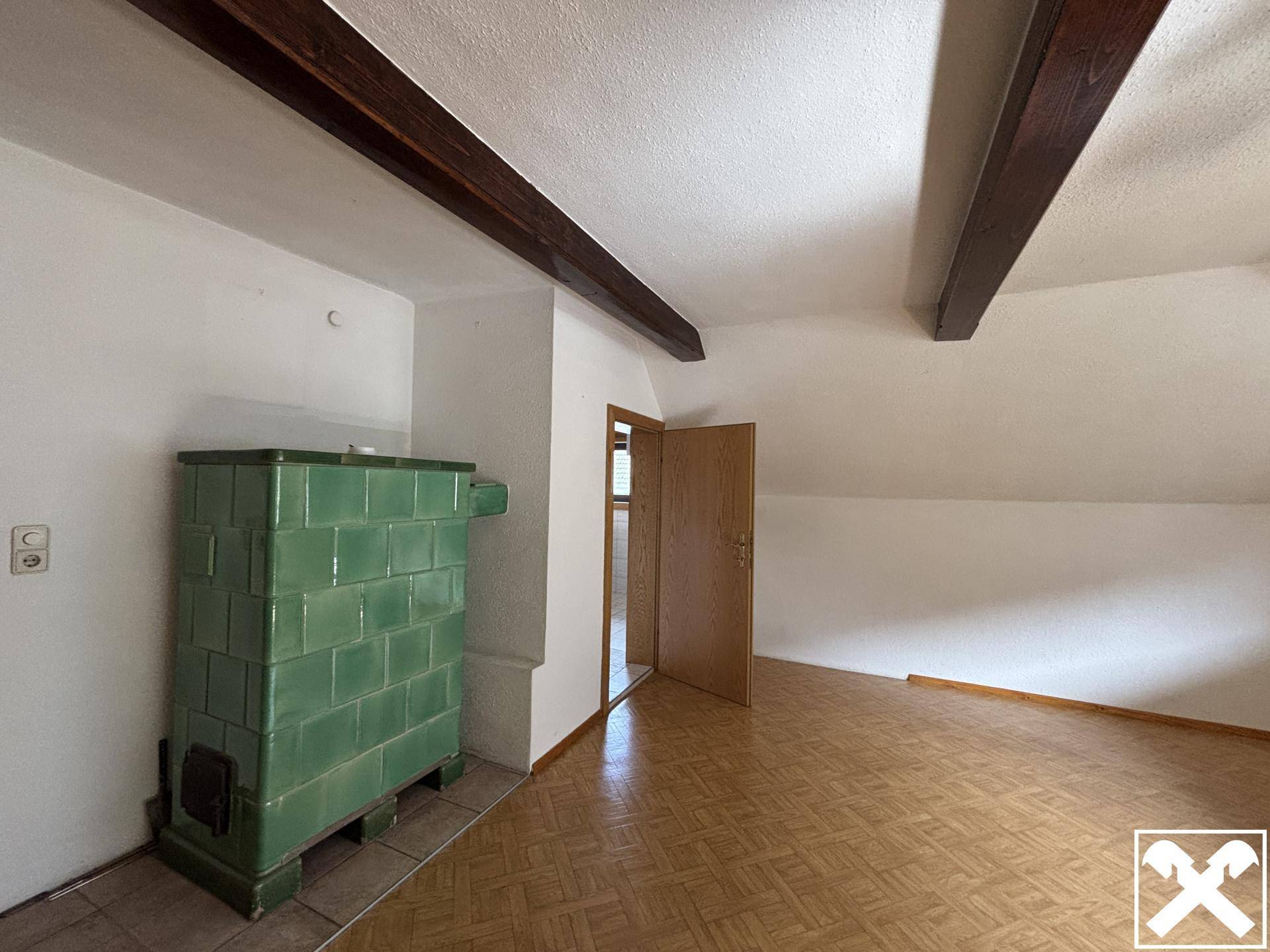 Helles Wohnzimmer mit grünem Kachelofen, Holzbalken und Dachschrägen.