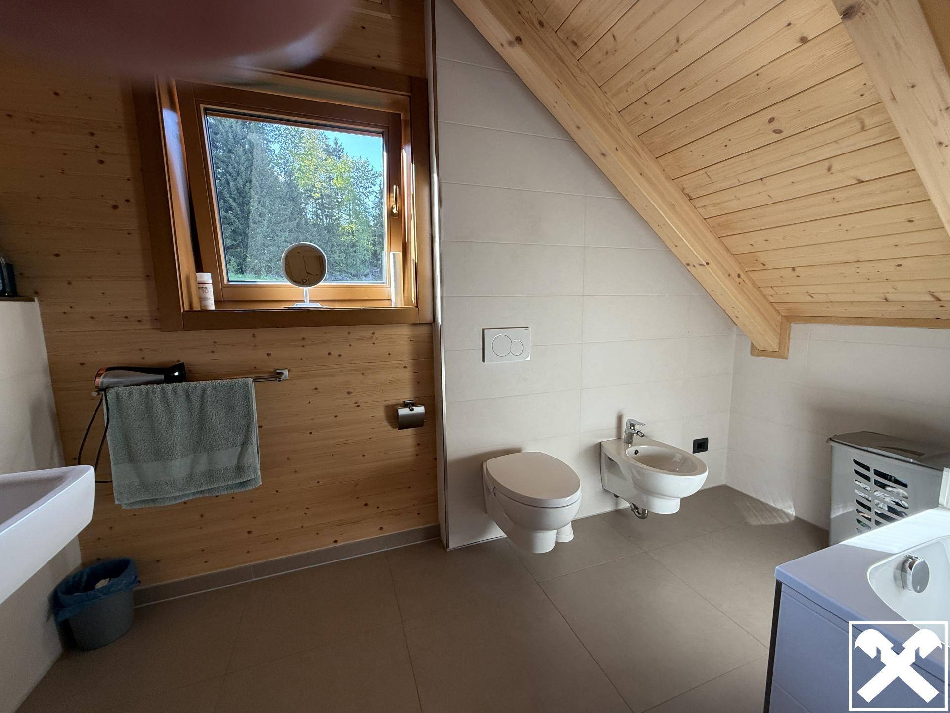 Geräumiges Badezimmer im Dachgeschoss mit WC, Bidet und Fenster mit Waldblick.