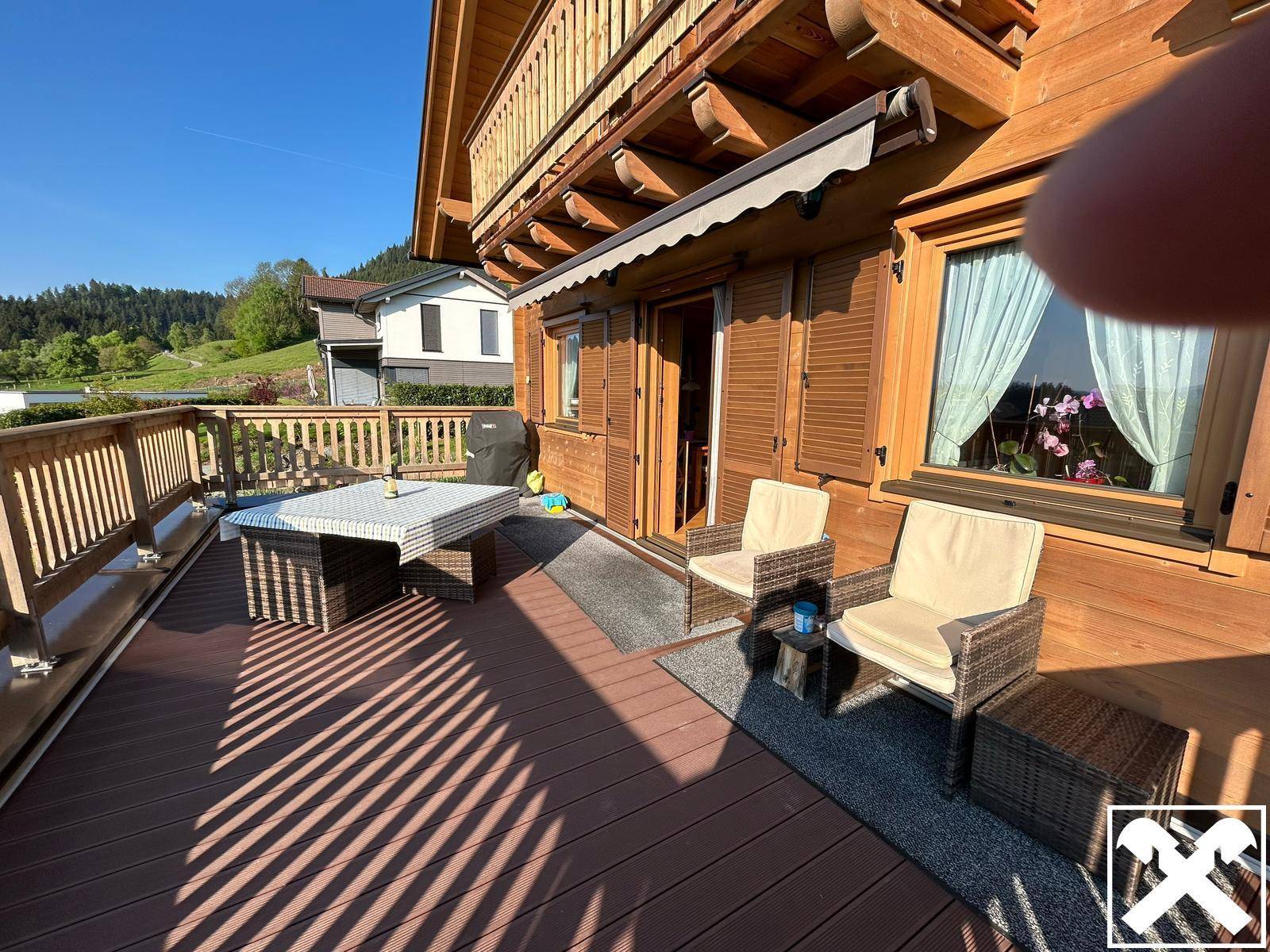 Sonnige Terrasse mit Holzboden und Gartenmöbeln vor einem traditionellen Holzhaus mit Bergblick.