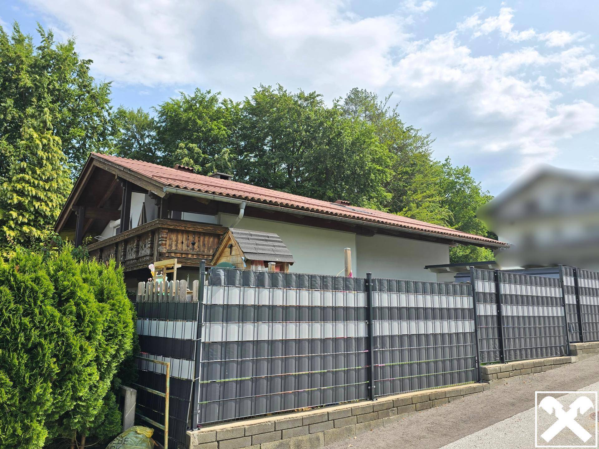 Seitenansicht des Hauses mit Holzbalkon, modernem Zaun und üppiger Vegetation.