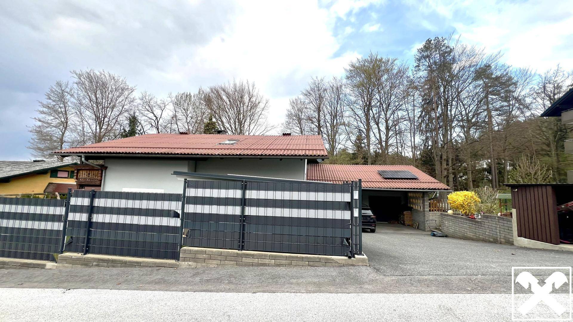 Ansicht des Hauses von der Straße mit modernem Zaun, Einfahrt und Bäumen im Hintergrund.